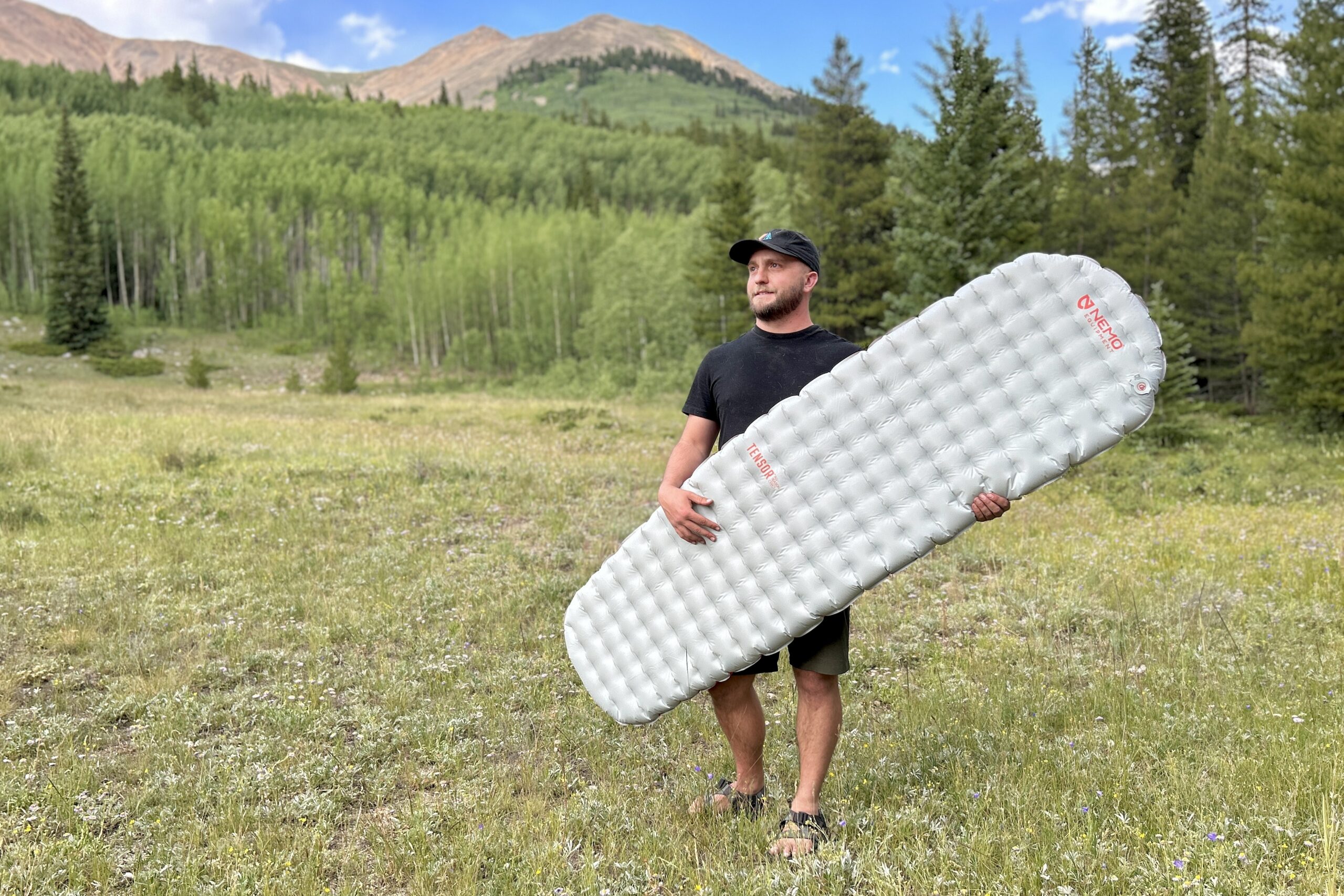 In a large field with mountains in the background, a man holds an inflatable grey sleeping pad.