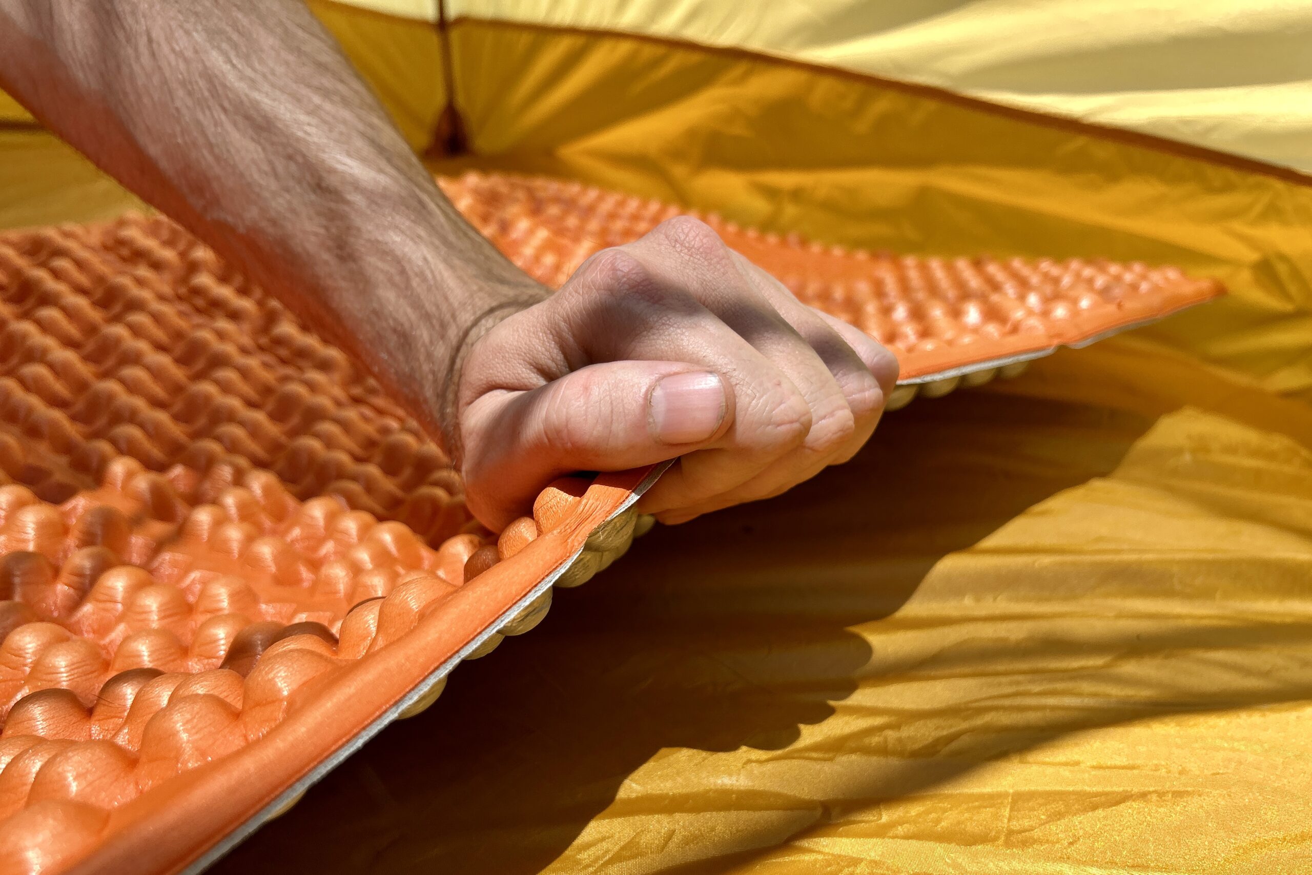 A person lifts an orange foam sleeping pad in a tent.