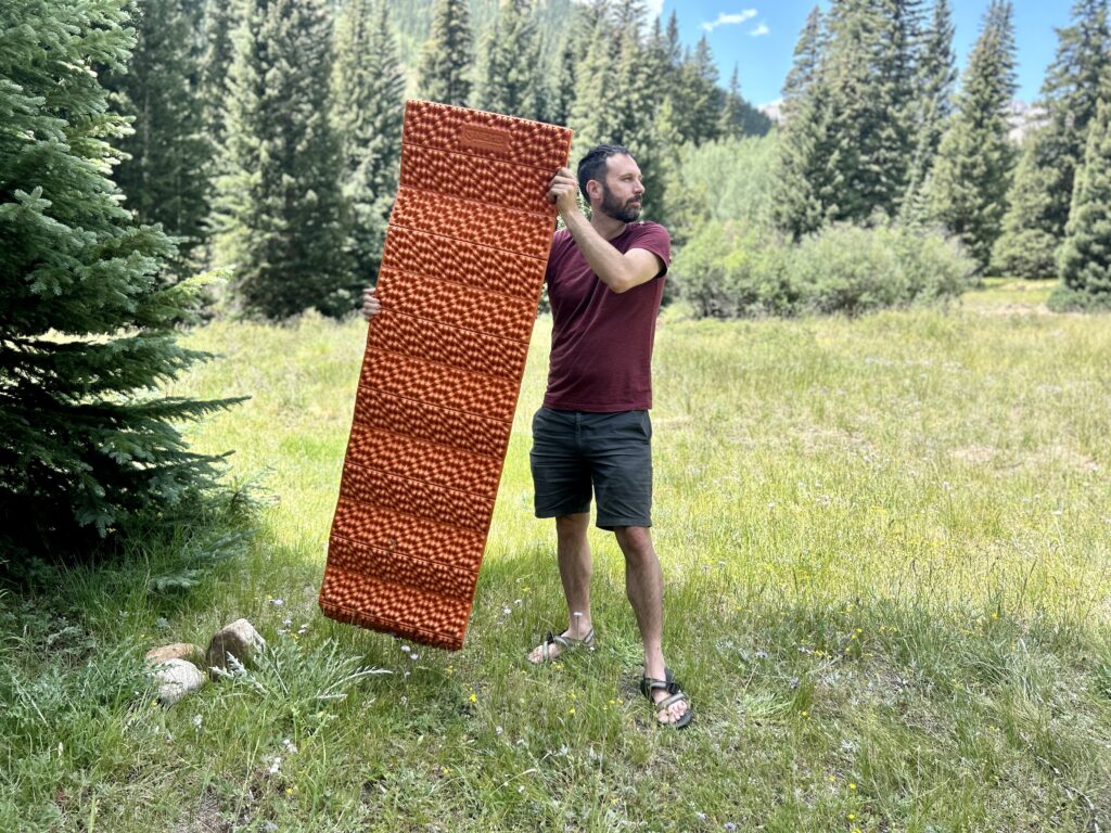 A man holds an orange mat in a grassy area in the woods.