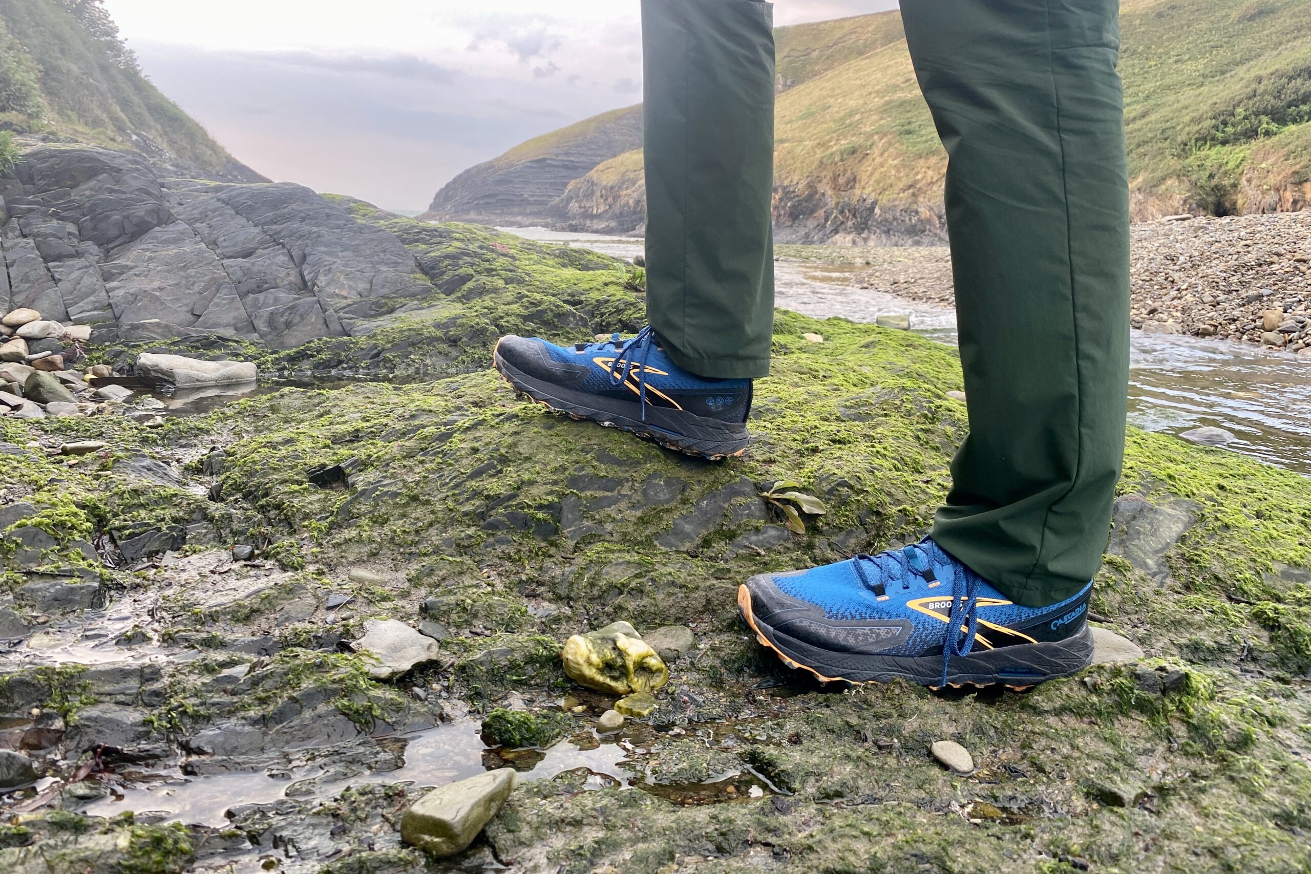 The lower legs of a man with a pair of blue trail running shoes in the foreground with large, oceanside cliffs in the background on an overcast day at sunset.