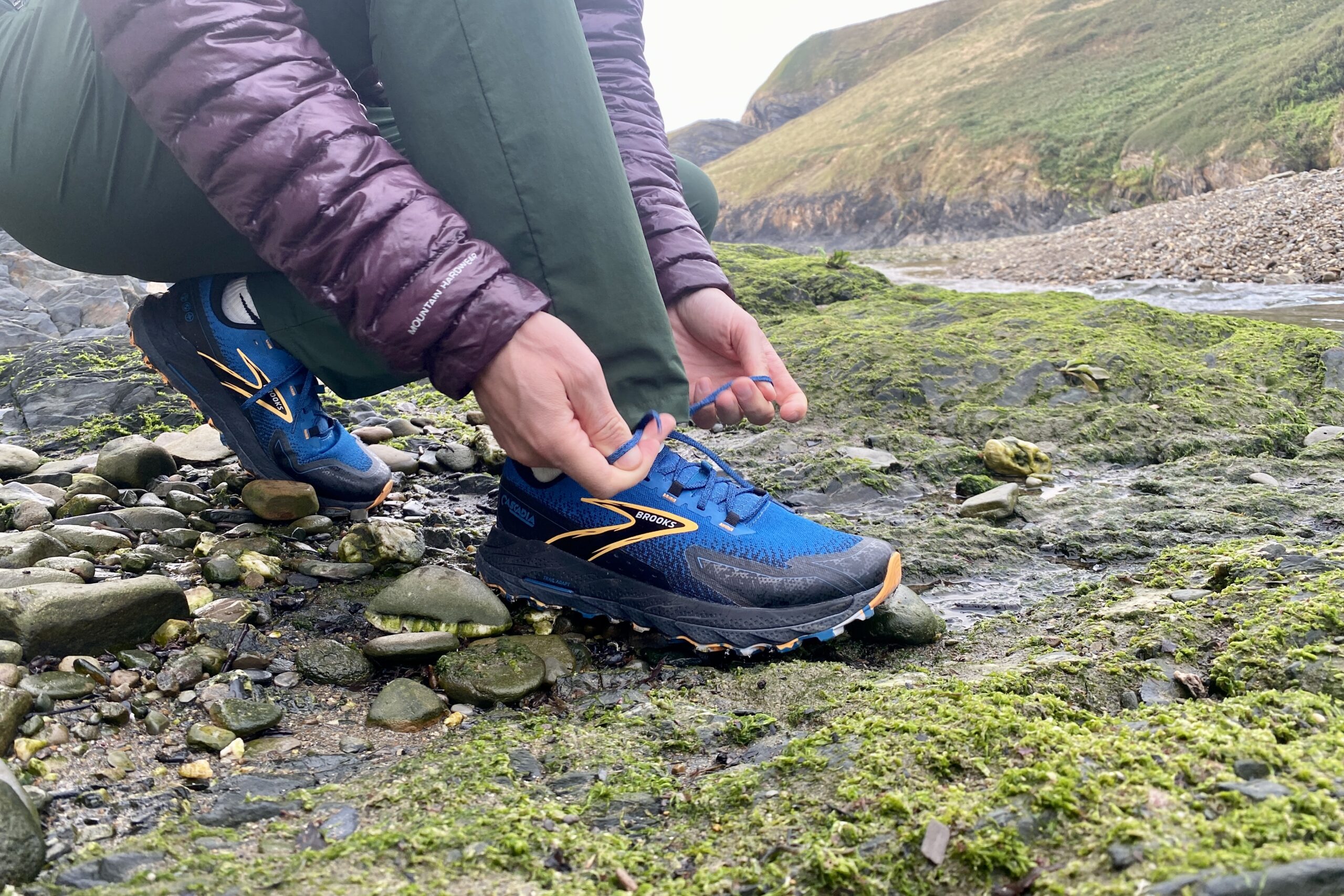 A man bends down to tie a trail running shoe with rock, moss, and a creek in the background.