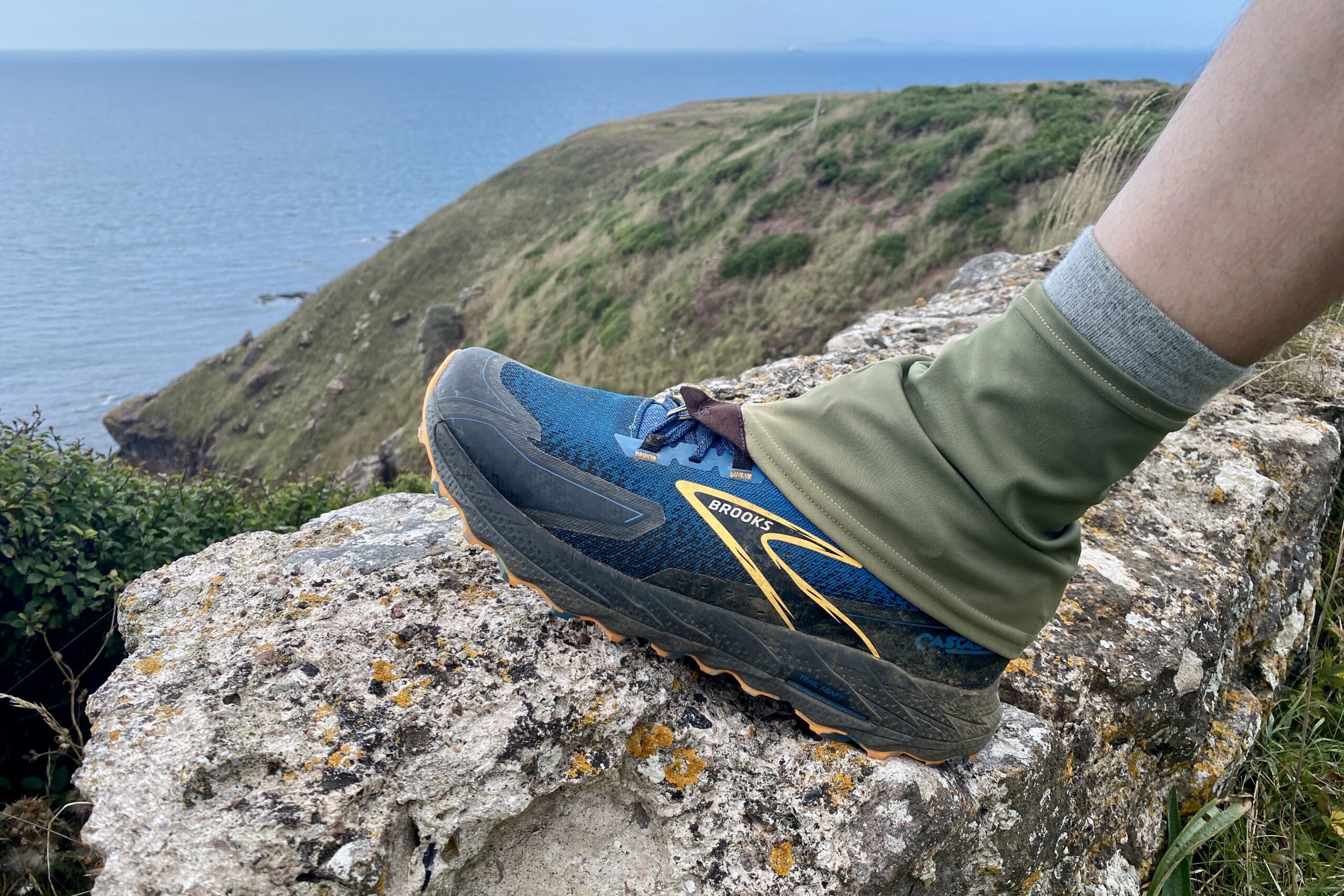 The left foot of a man stepping on a rock while wearing a pair of trail running shoes with a pair of low gaiters and an ocean view in the background.