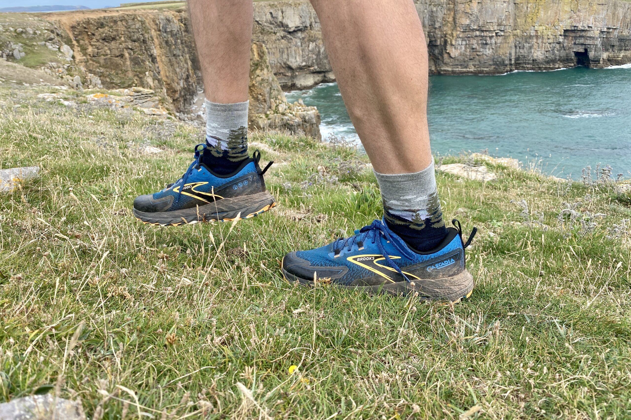 A man lifts his right foot while wearing a pair of trail running shoes with oceanside cliffs and water in the background.