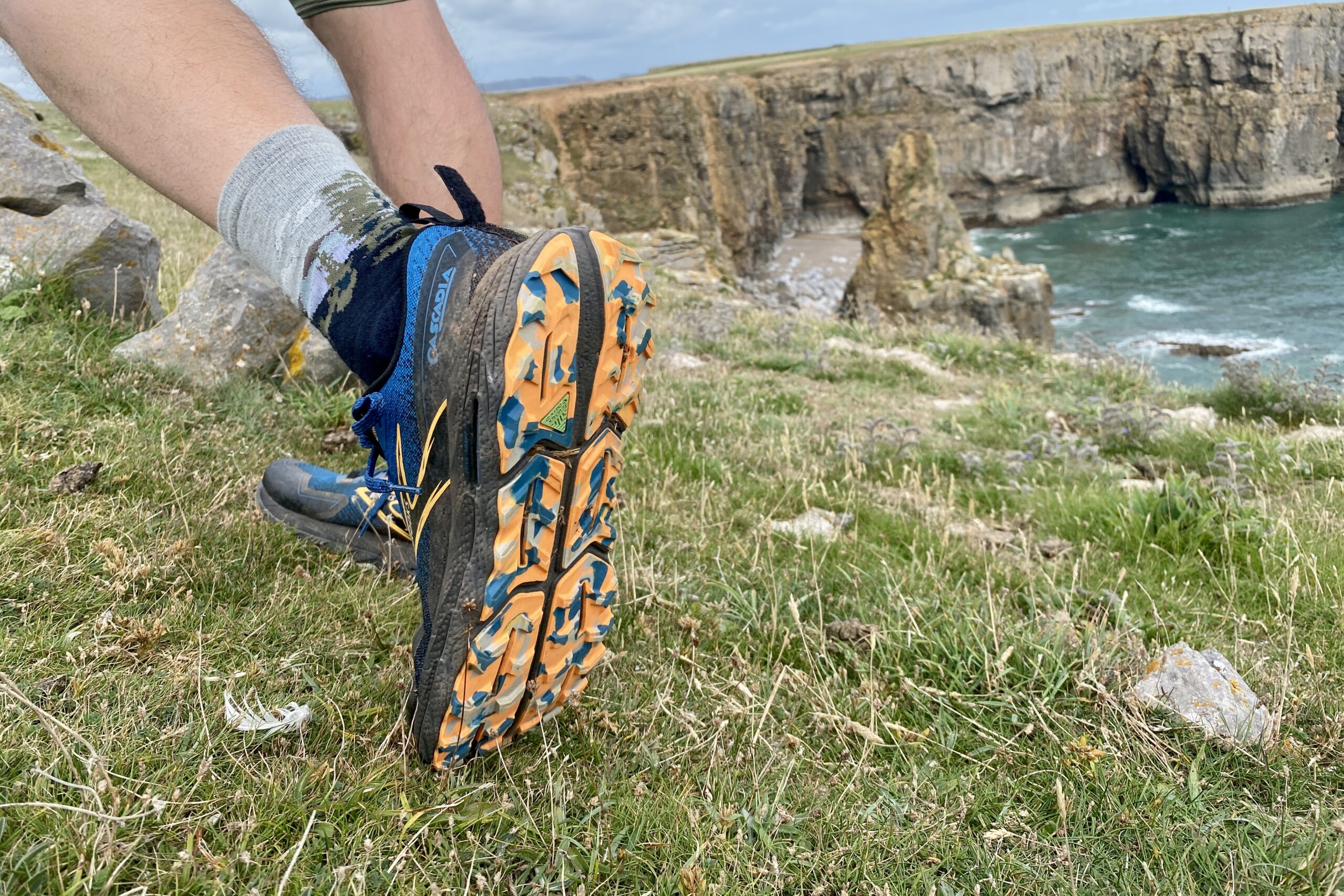 A man lifts his left foot showing the tread on the bottom of a pair of trail running shoes with oceanside cliffs and water in the background.