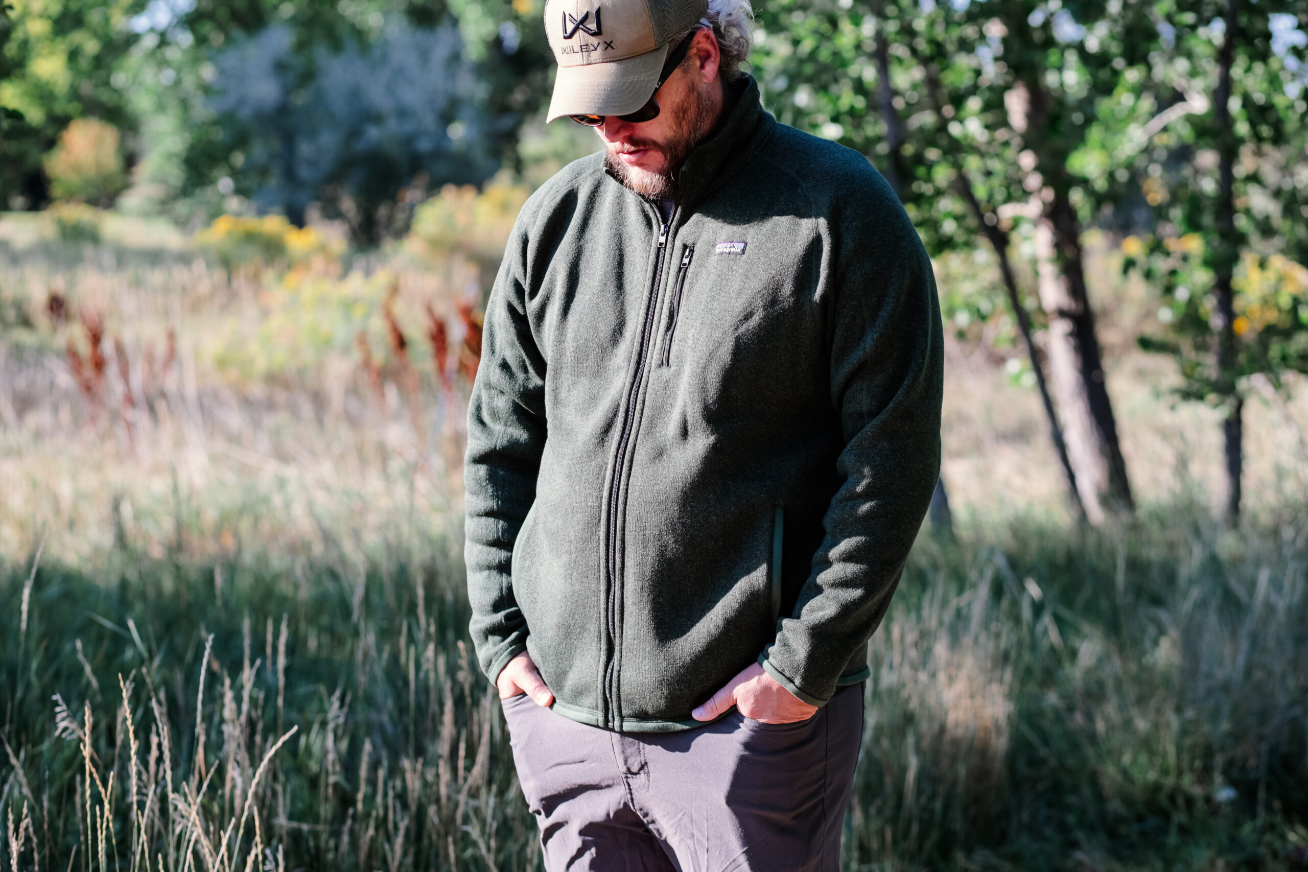 A man with hands in pockets wearing a green hat and fleece.