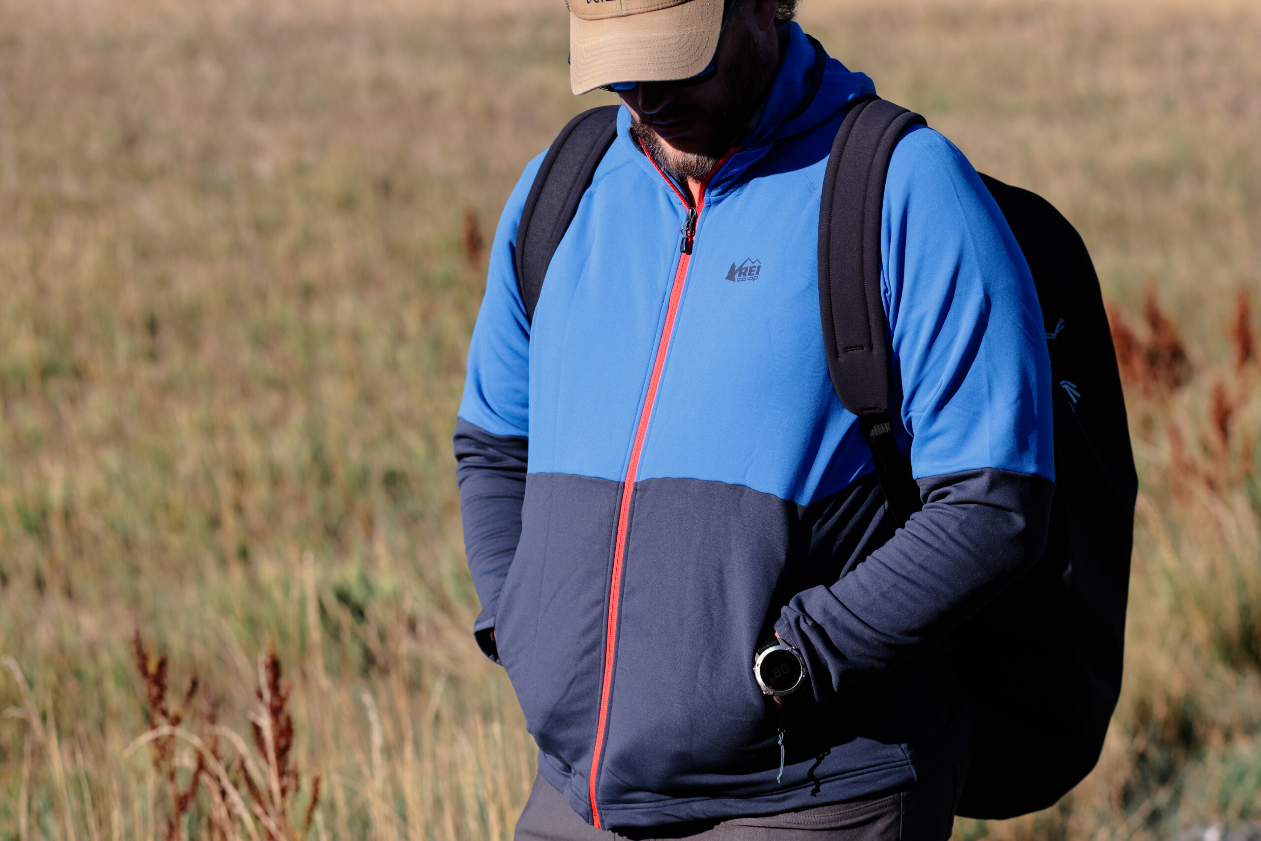 A man walks down a trail with a pack and fleece looking at the ground with hands in pockets.