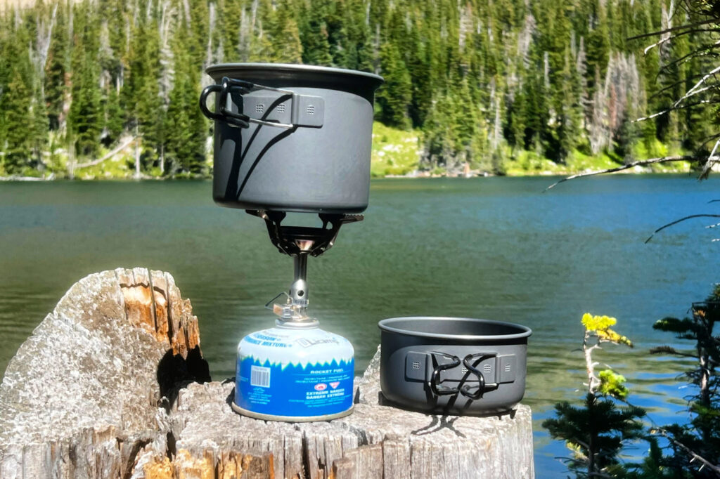 A backpacking stove with a pot on top sits on a stump in front of a mountain lake on a sunny day.