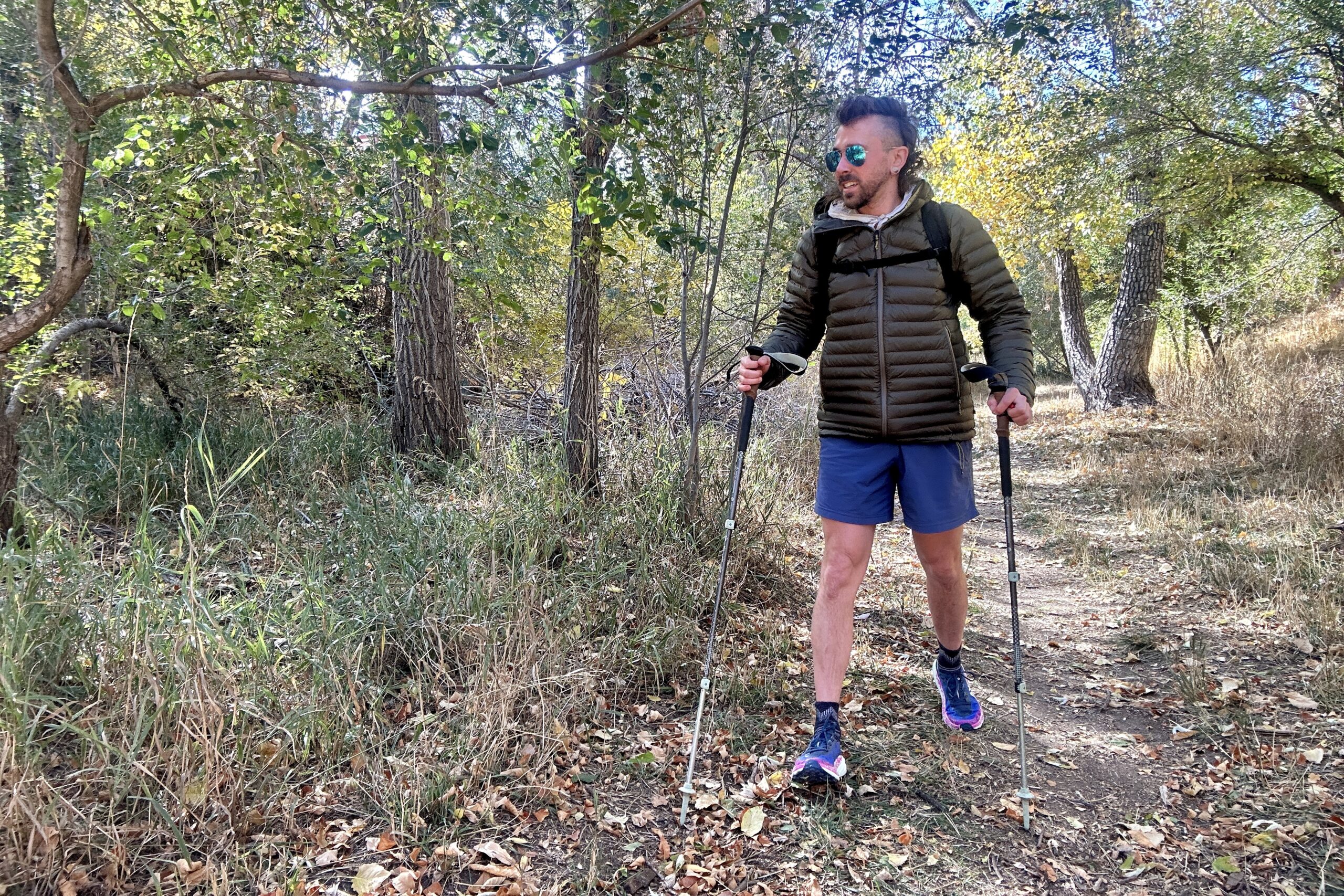 A man walking through a forest with a backpack and hiking poles and a puffy jacket on.