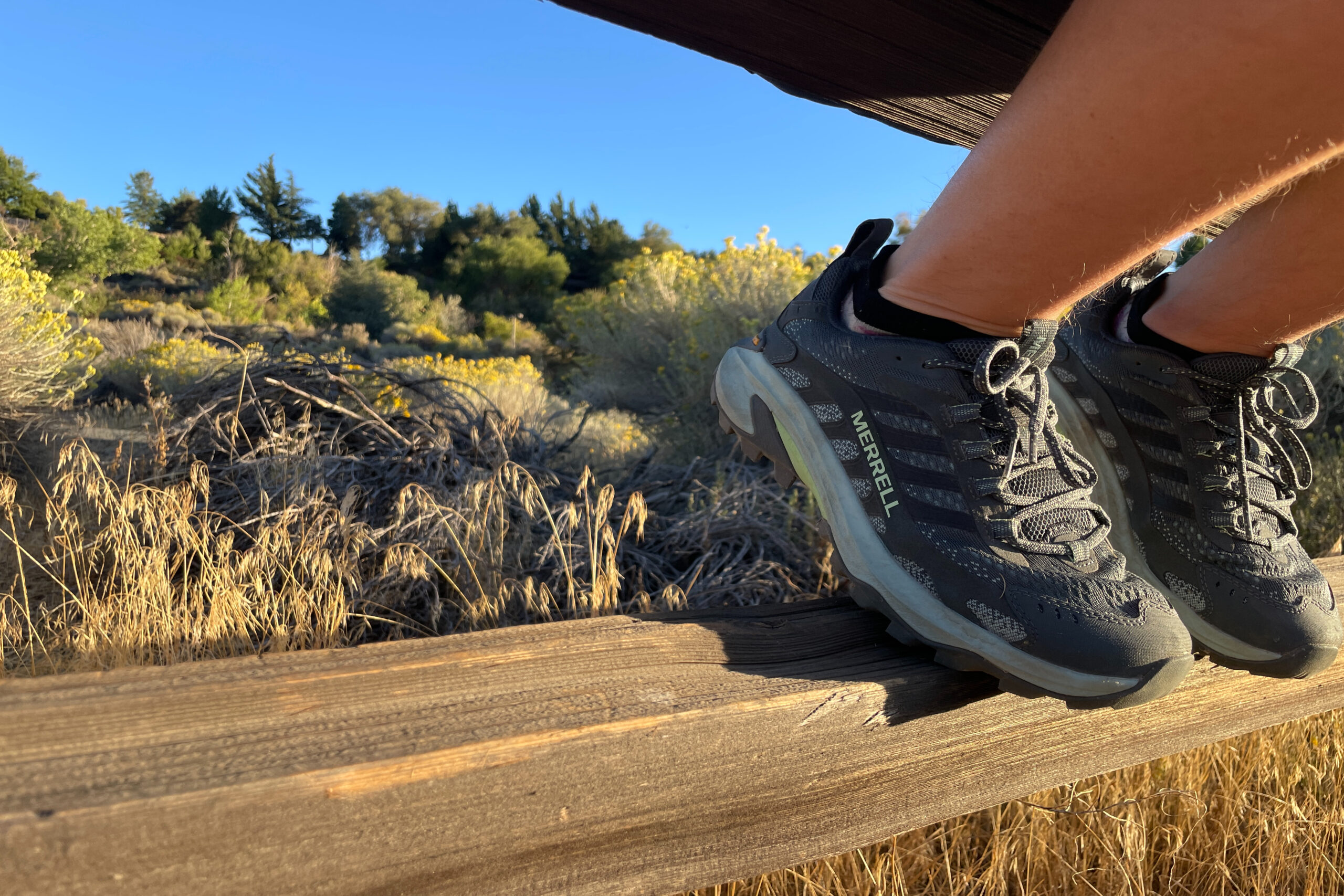 a picture of a women's feet resting on a rung of a wooden fence in the desert. She is wearing the Merrell Moab Speed 2 hiking shoes