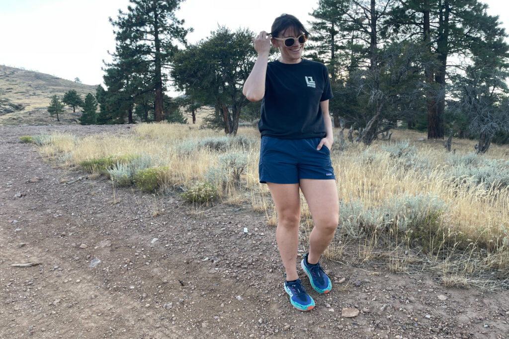 a women smiling and putting her hair behind her ear with the other hand in her pocket of the patagonia mult trails shorts while on a dirt road in next to sage brush and ponderosas