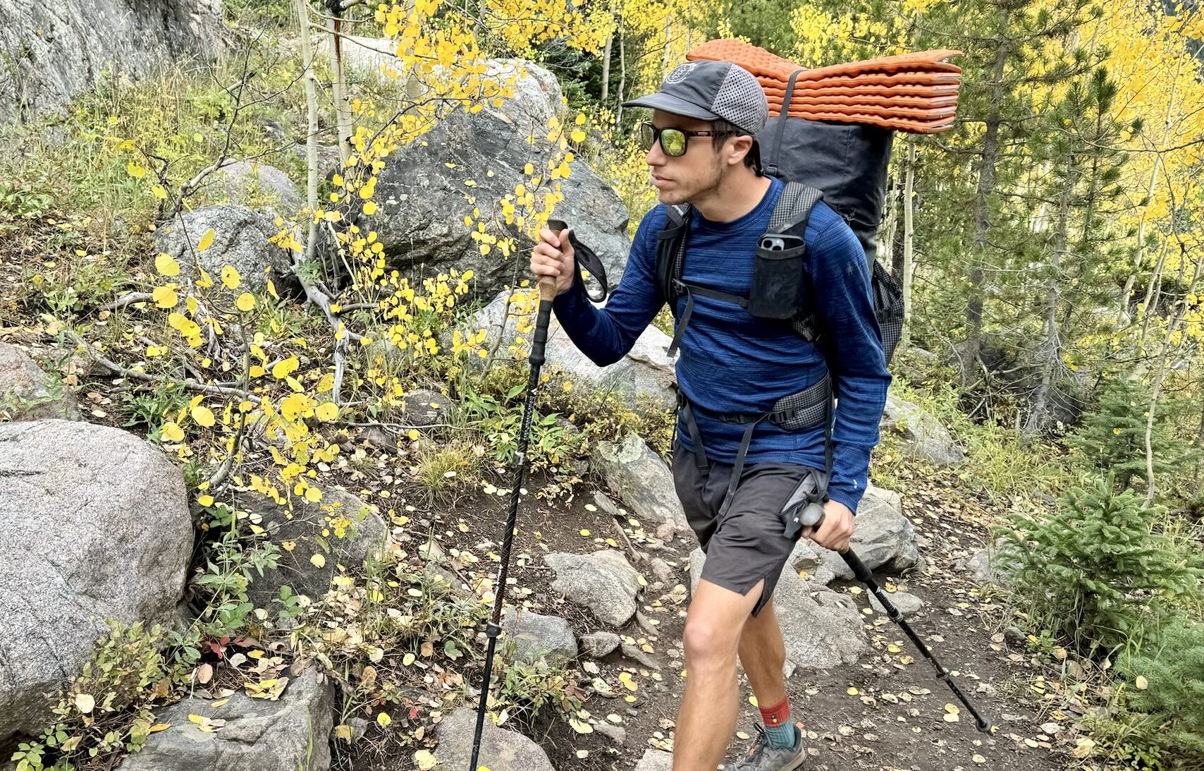 A hiker walks through the woods with trekking poles and a pack while wearing the Smartwool Classic Thermal base layer. Trees and rocks are in the background.