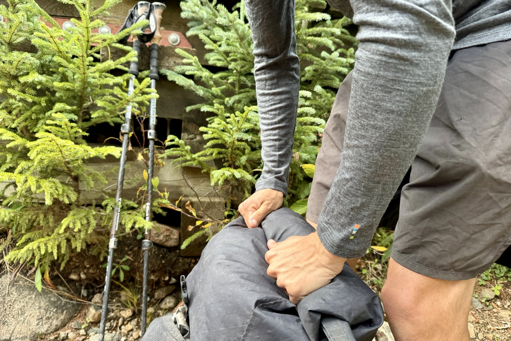 A hiker rolls up his backpack while wearing the Smartwool Classic All-Season base layer. Trees and hiking poles are in the background.