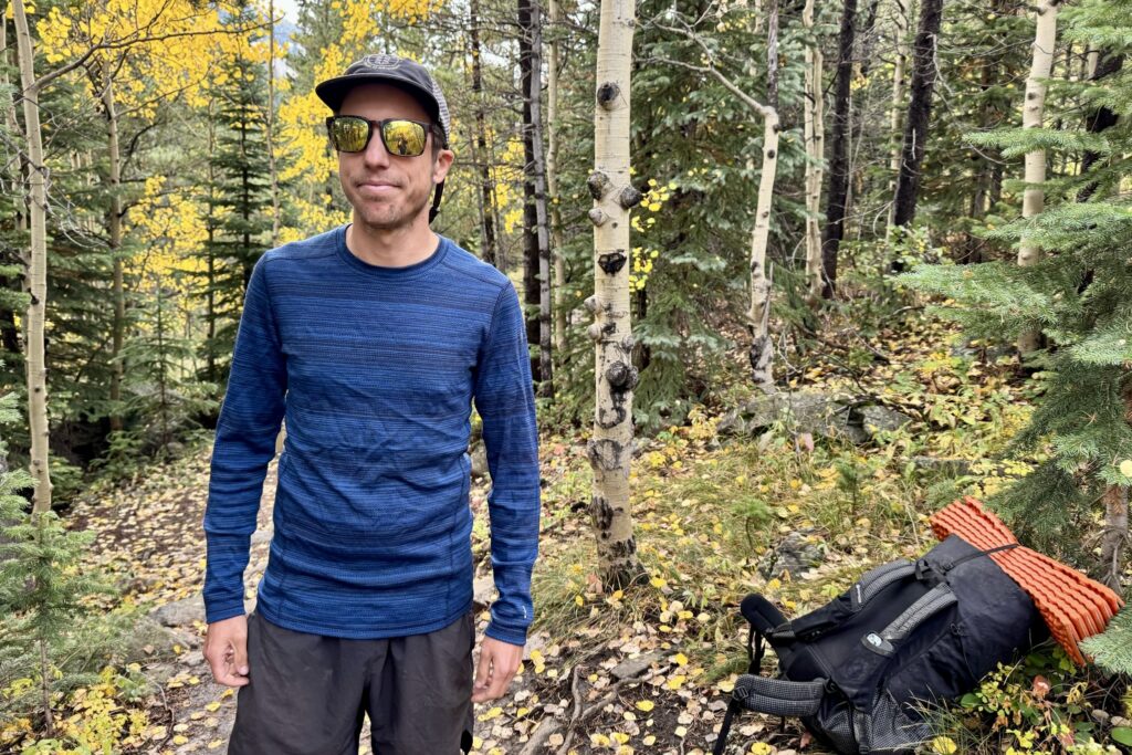 A hiker stands in the woods wearing the Smartwool Classic Thermal base layer. A backpack and trees are in the background.