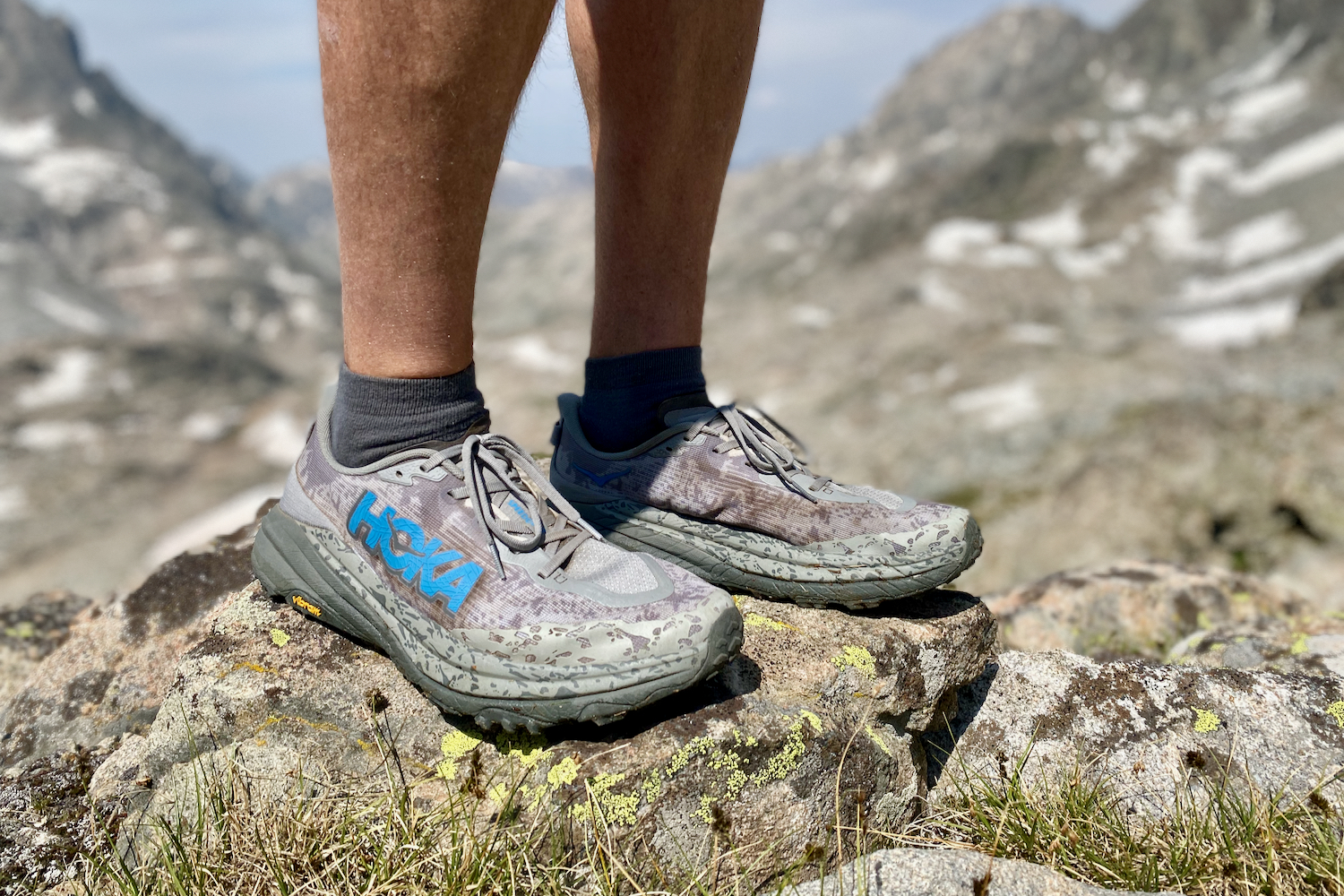 Hey side facing view of the HOKA Speedgoat 6 trail running shoes with a hiker, wearing them at high altitude with snowy peaks in the background.