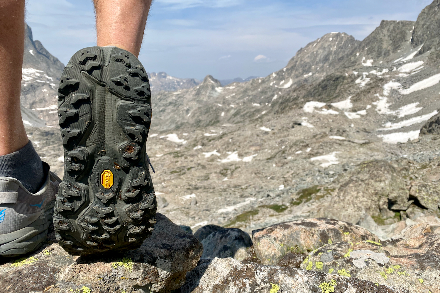 A hiker wearing the HOKA Speedgoat 6 and lifting up his foot to show the traction on the bottom of the shoe, with distant mountain peaks in the background.