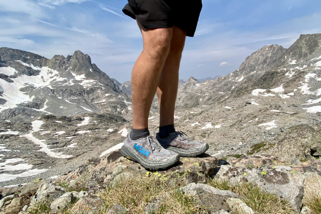 A hiker standing on a rock wearing the HOKA Speedgoat 6 trail running shoes with a beautiful view of craggy mountain peaks in the background at high altitude.