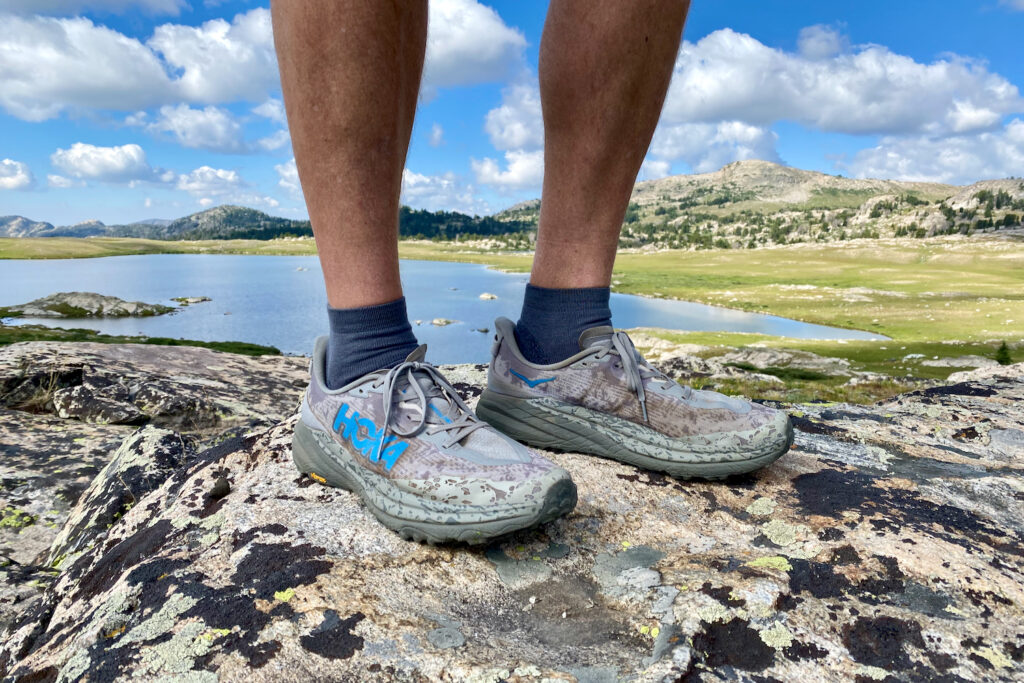 The side facing view of the HOKA Speedgoat 6 trail running shoes with a lake and pretty wilderness scenery in the background.