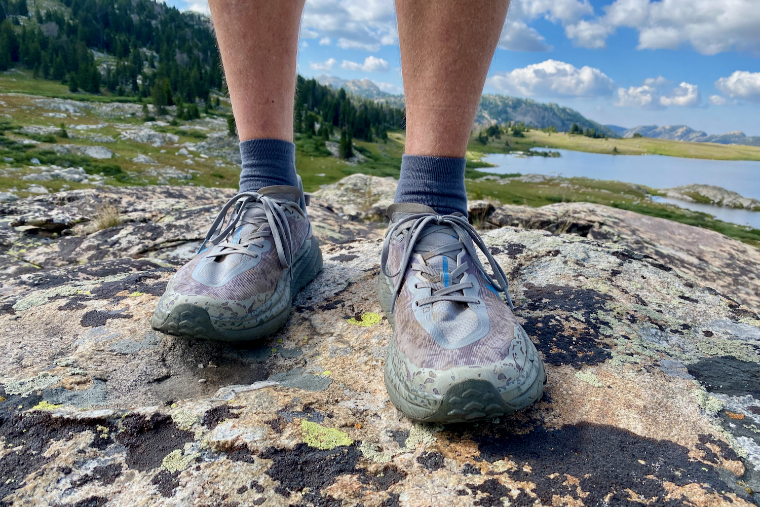 A forward facing view of the HOKA Speedgoat 6 hiking shoes while on a hikers feet and facing the camera with a lake and trees in the background.