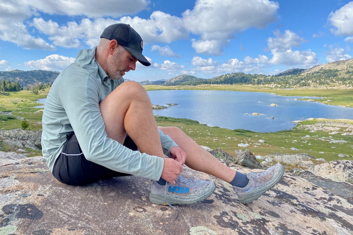 A hiker sitting on a rock, tying the HOKA Speedgoat 6 shoes. The hiker is full body in the image, and there is a lake and mountains in the background.