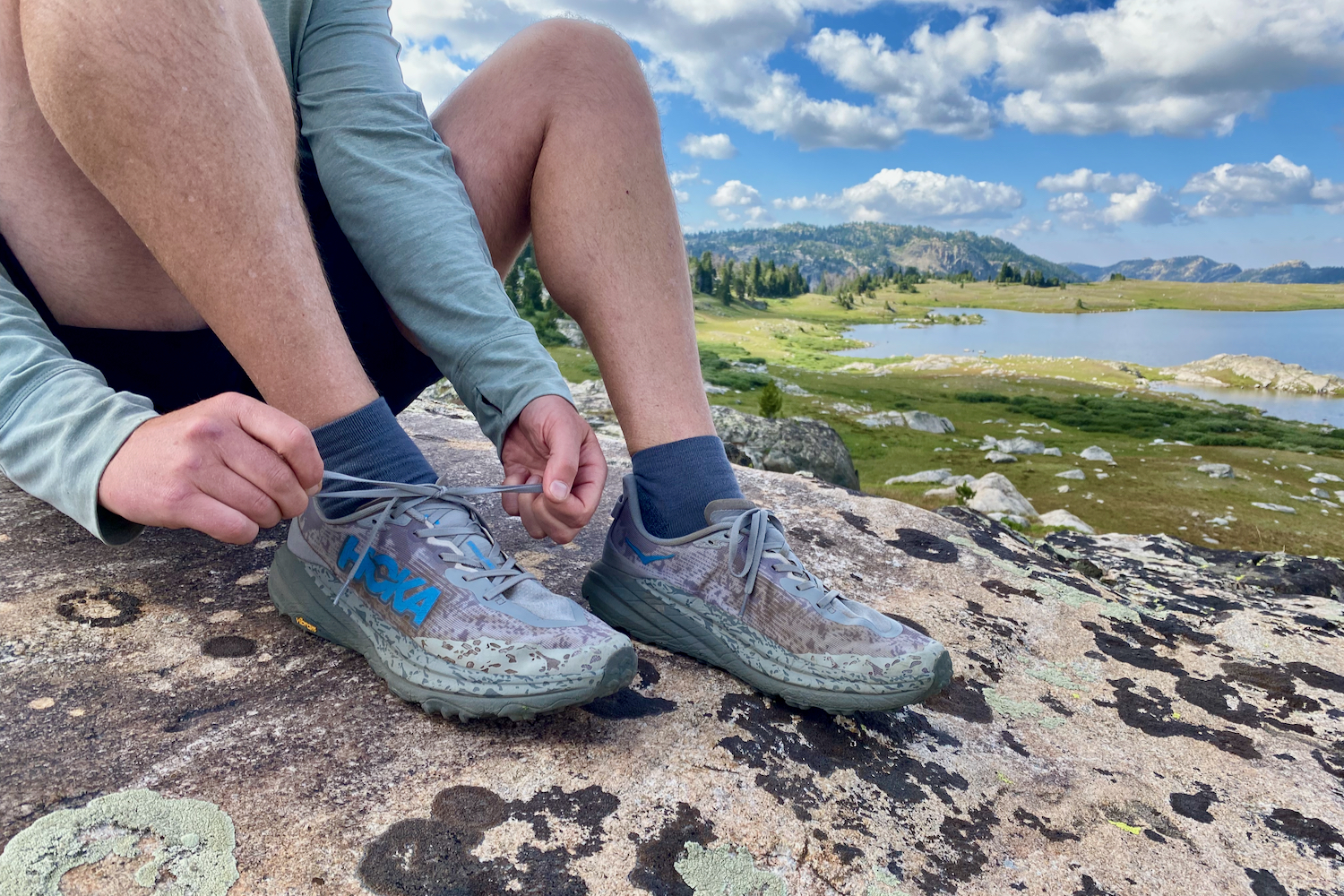 A hiker tying the HOKA Speedgoat 6 shoes while sitting on a rock with a lake and pretty mountains and clouds in the background.