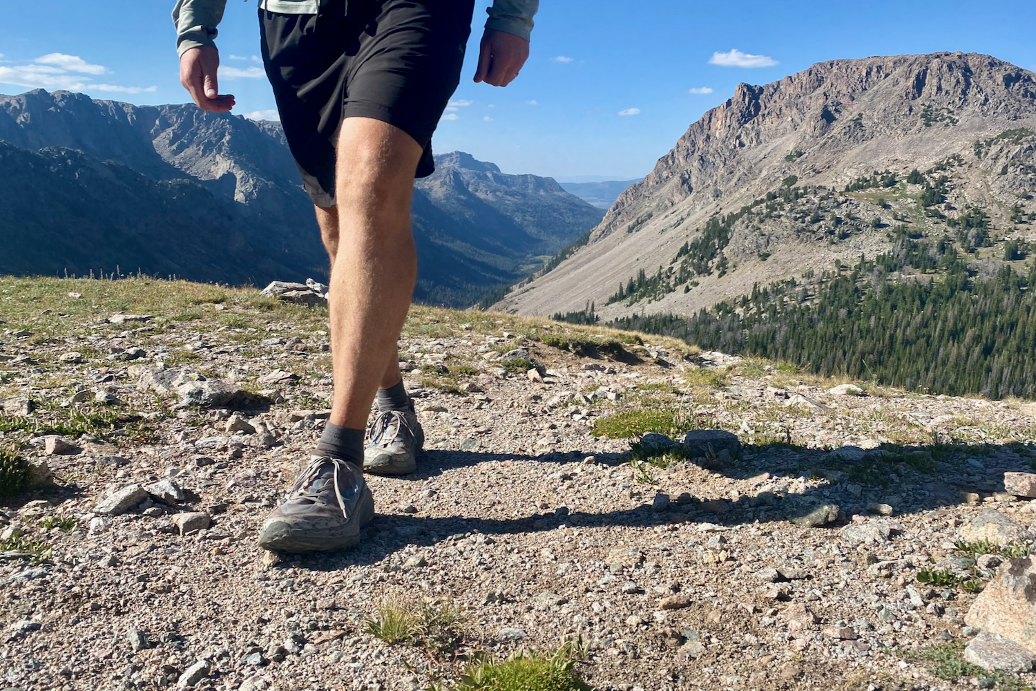 A hiker walking towards the camera down a trail with mountains in the background wearing the HOKA Speedgoat 6