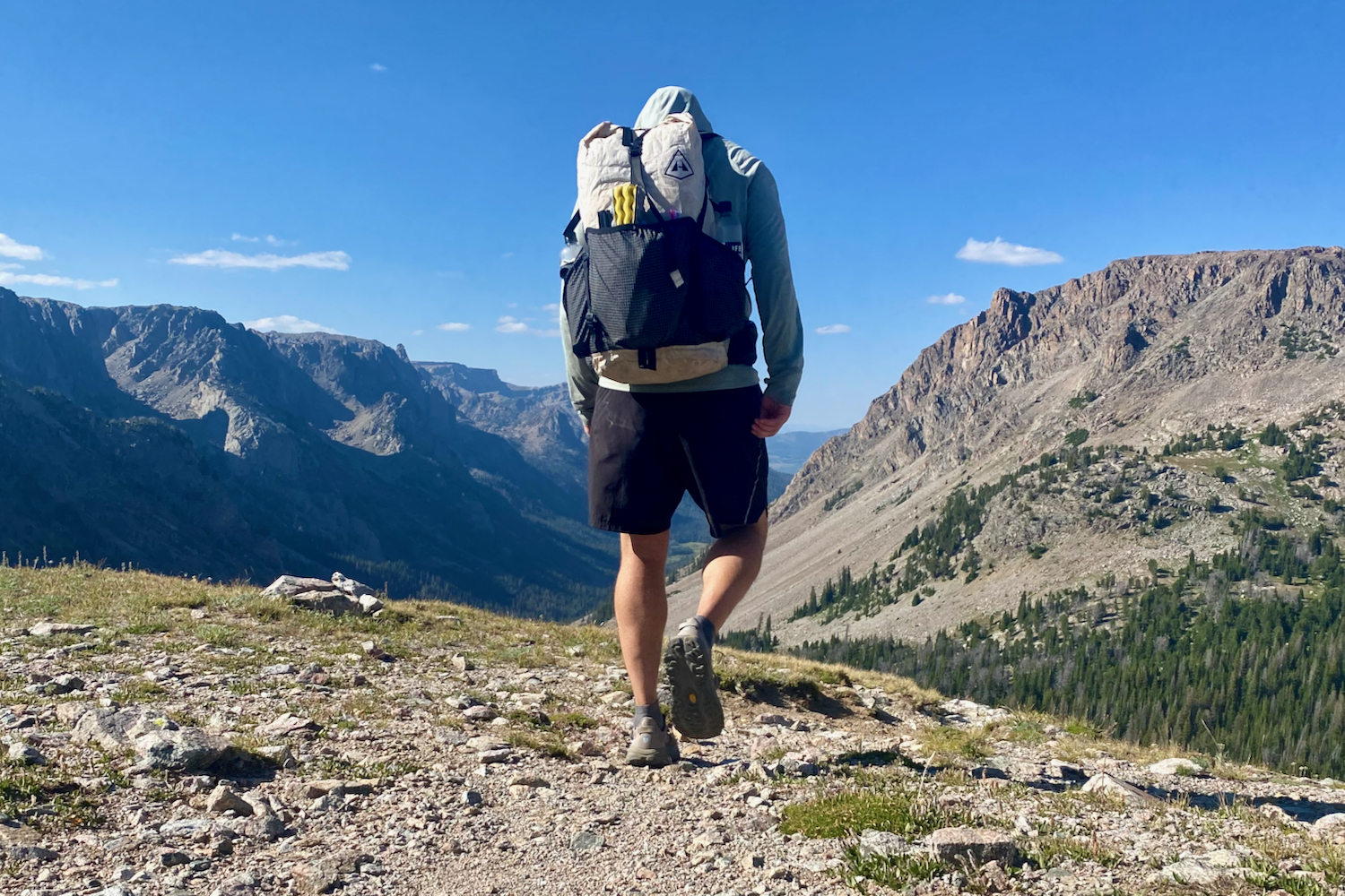 A hiker walking down a trail with mountains in the background and the HOKA Speedgoat 6 hiking shoes on his feet