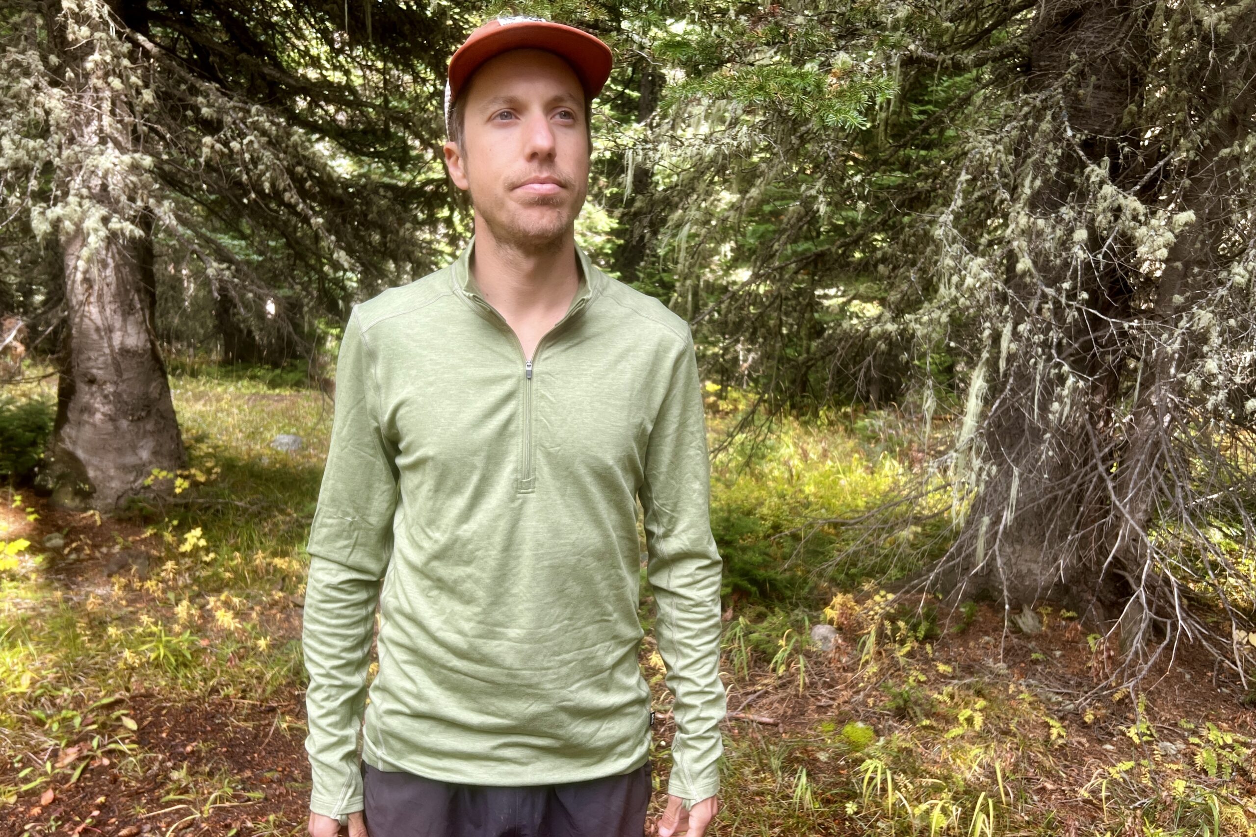 A hiker stands in the woods wearing the REI Midweight Half Zip with the zipper half way up. Trees are in the background.