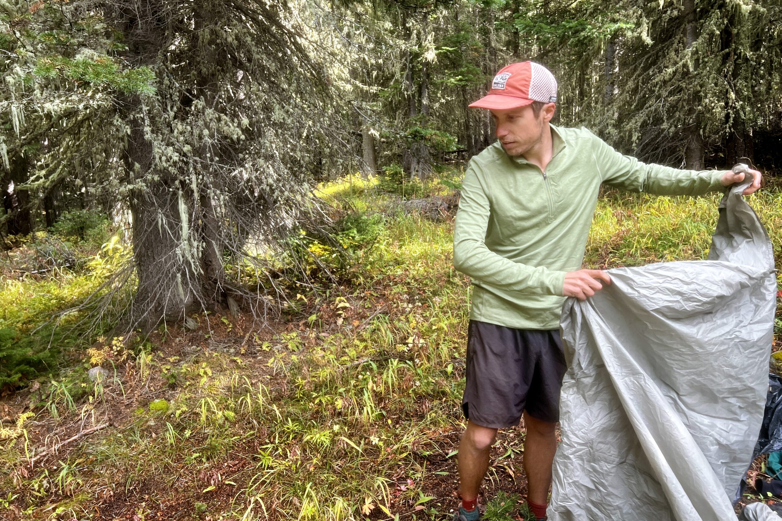 A hiker prepares to pitch his tent while wearing the REI Midweight Half Zip base layer. Trees are in the background.