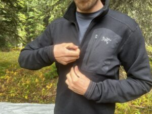 A hiker zips up the neck on the Arc'teryx Rho Heavyweight Zip Neck base layer. Trees are in the background.