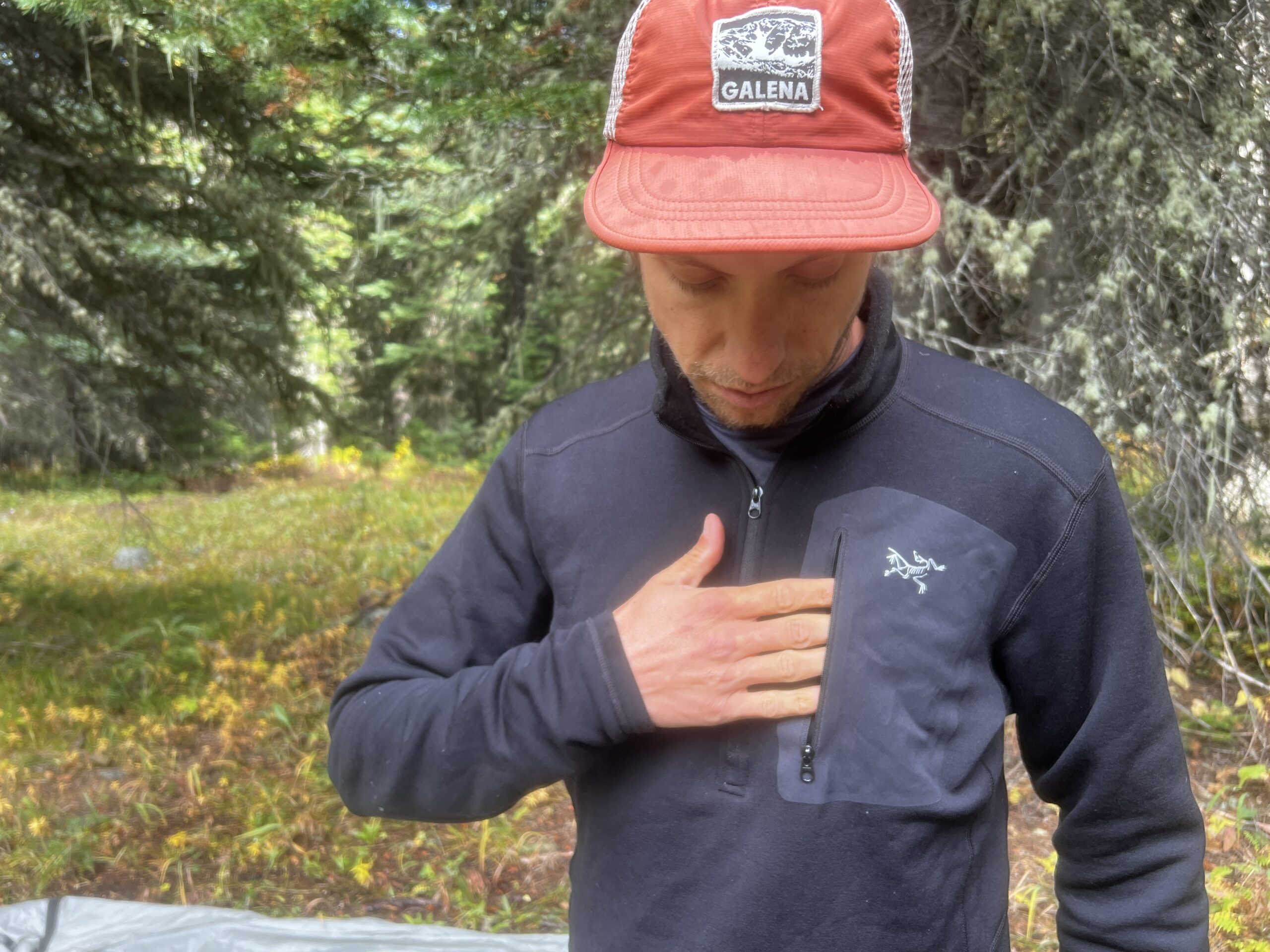 a hiker slides his hand into the breast pocket of the Arc'teryx Rho Heavyweight Zip Neck base layer. Trees are in the background.