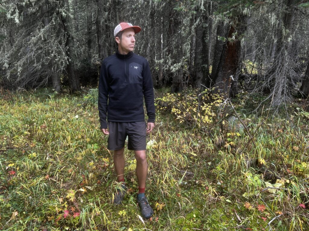 A hiker stands in the woods wearing the Arc'teryx Rho Heavyweight Zip Neck base layer. Trees are in the background.