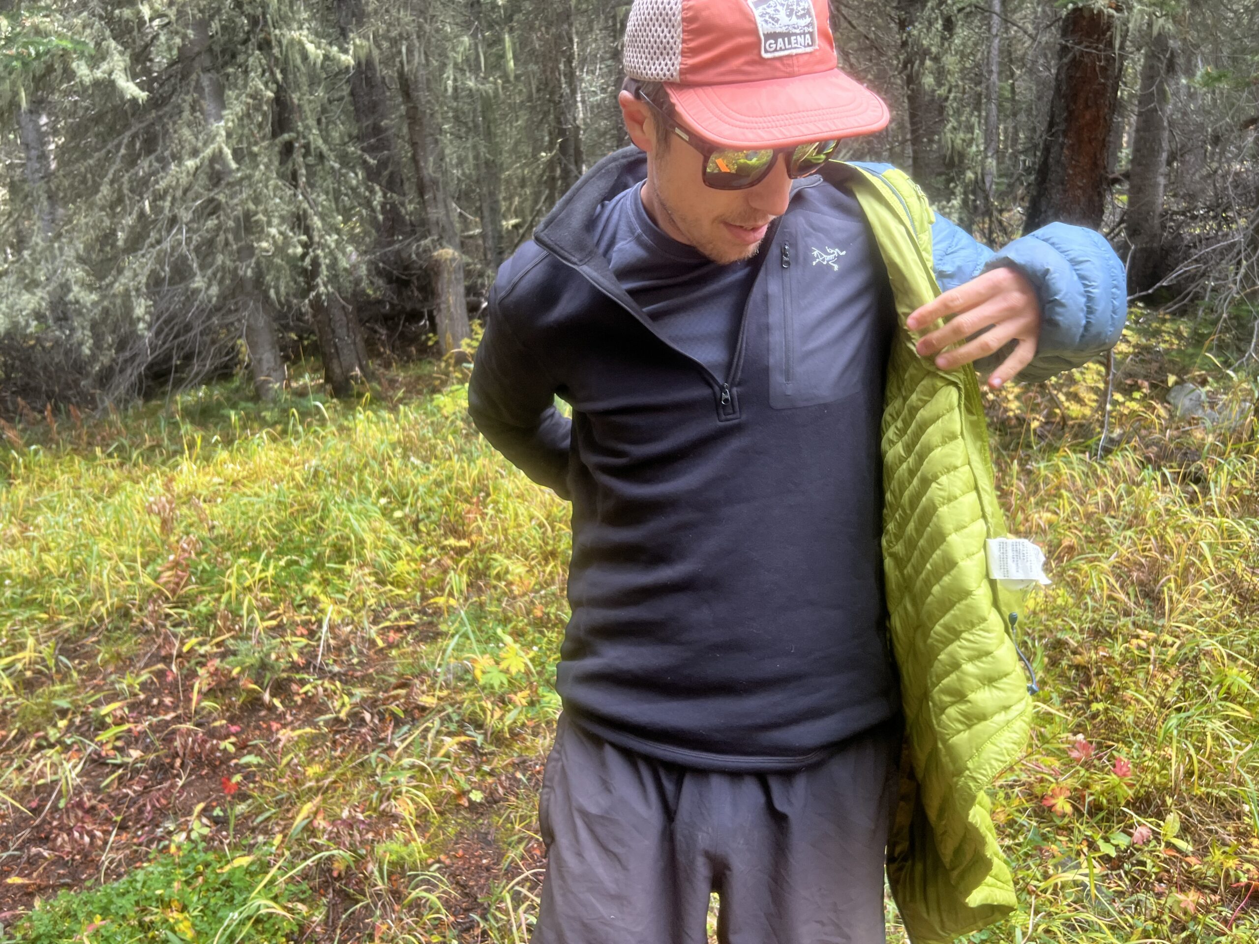 A hiker puts on a puffy jacket over top of the Arc'teryx Rho Heavyweight Zip Neck base layer. Trees are in the background.