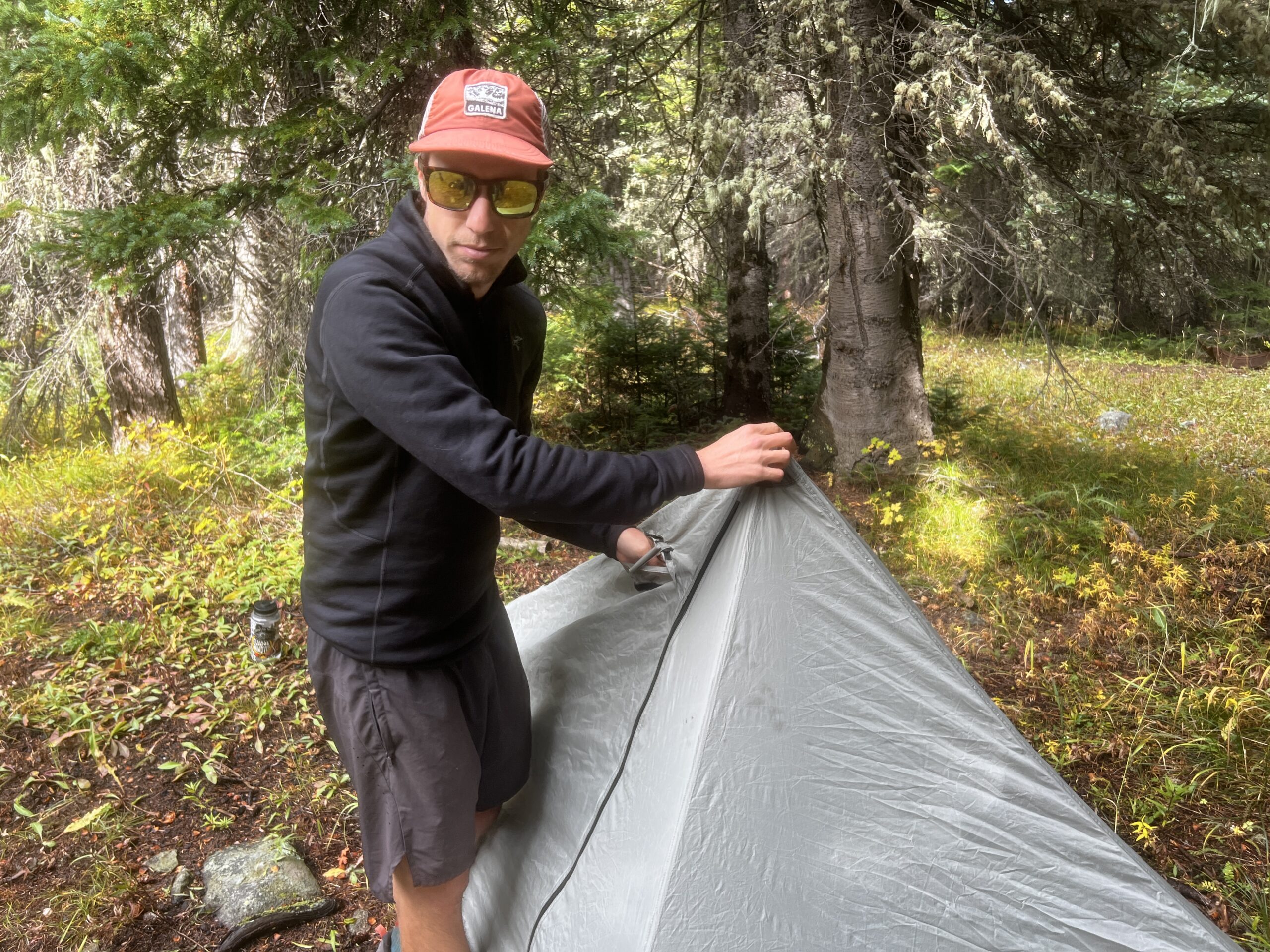 A hiker sets up his tent while wearing the Arc'teryx Rho Heavyweight Zip Neck base layer. Trees are in the background.