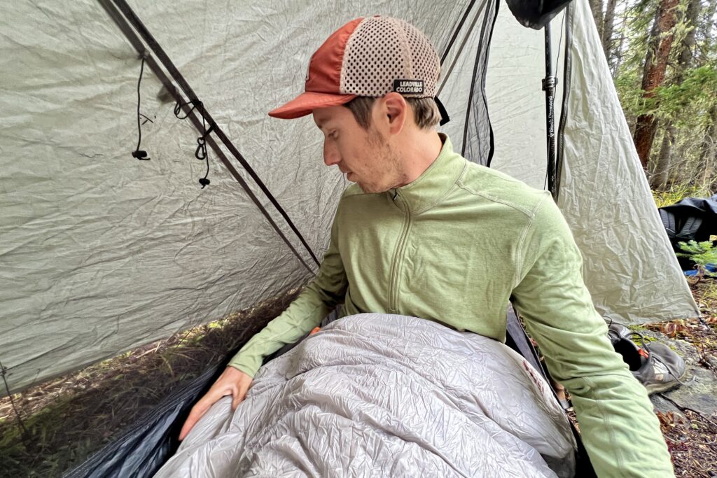 A hiker sits in his sleeping bag in a tent wearing the REI Midweight Half Zip base layer.