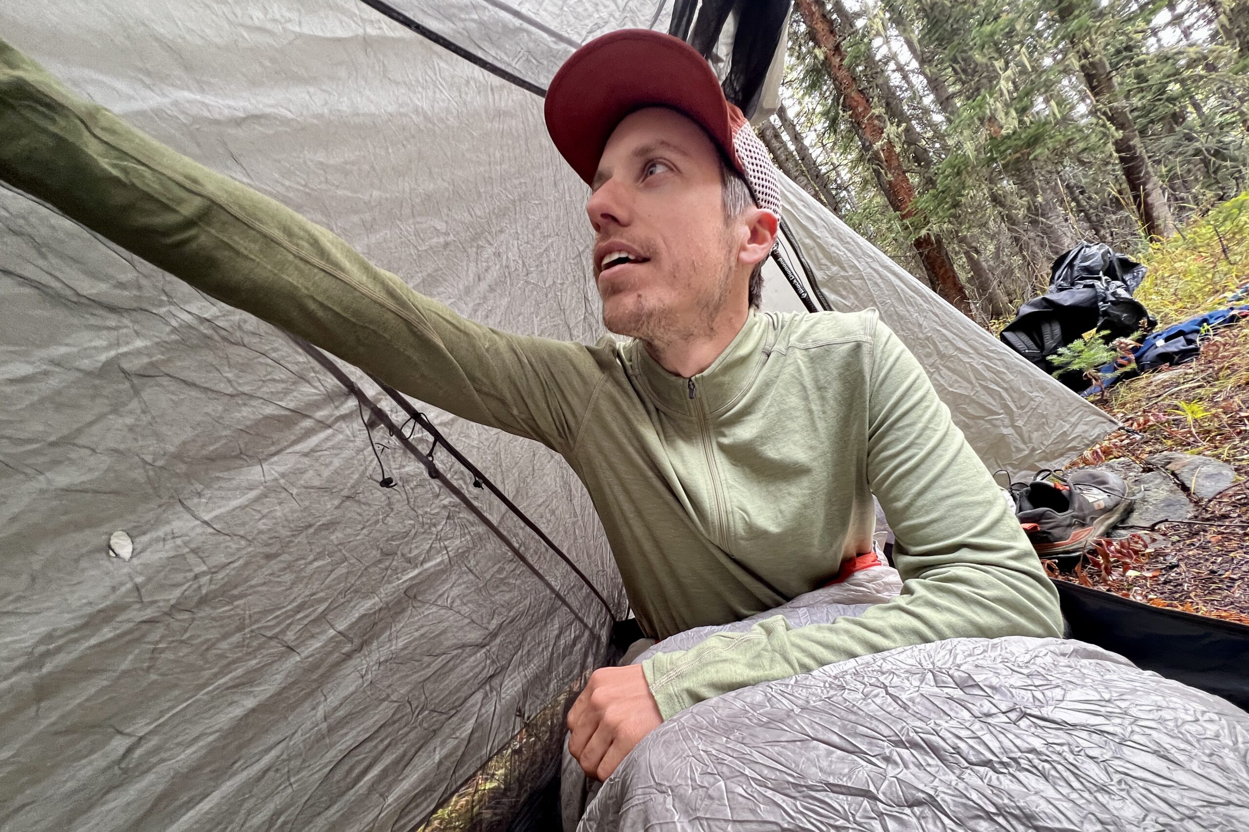 A hiker reaches up to adjust his tent while sitting in his sleeping back with the REI Midweight Half Zip. Trees are in the background.