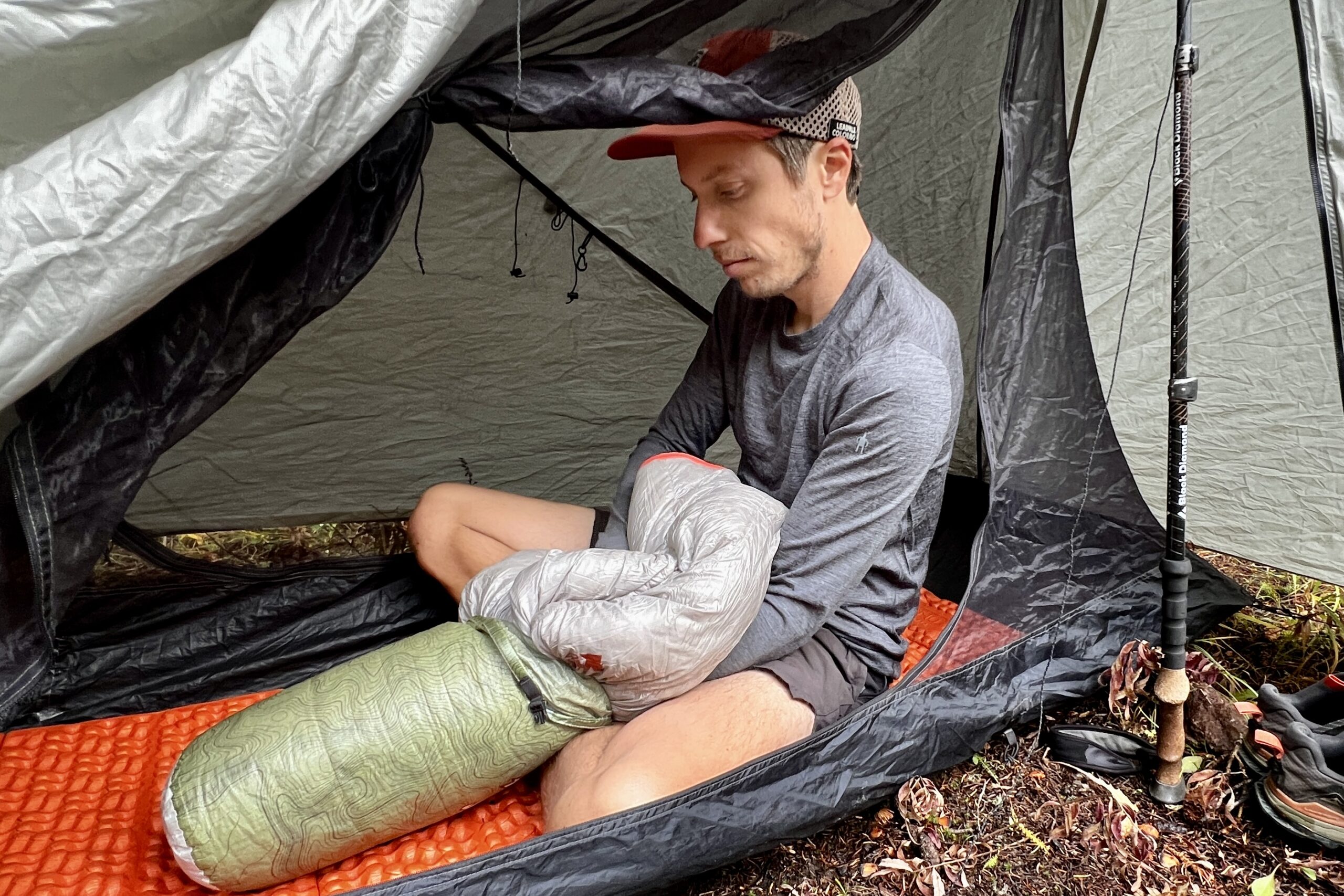 A hiker sits in his tent and stows his sleeping back away while wearing the Smartwool Classic All-Season base layer.