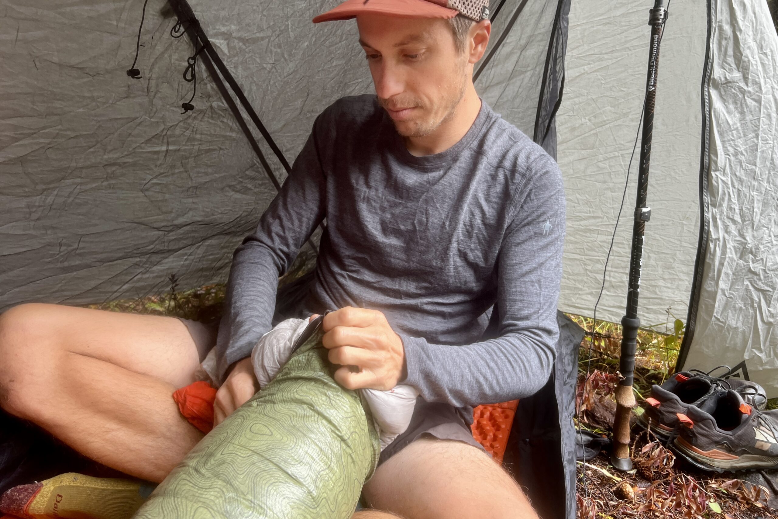 A hiker sits in his tent and stows his sleeping back away while wearing the Smartwool Classic All-Season base layer.