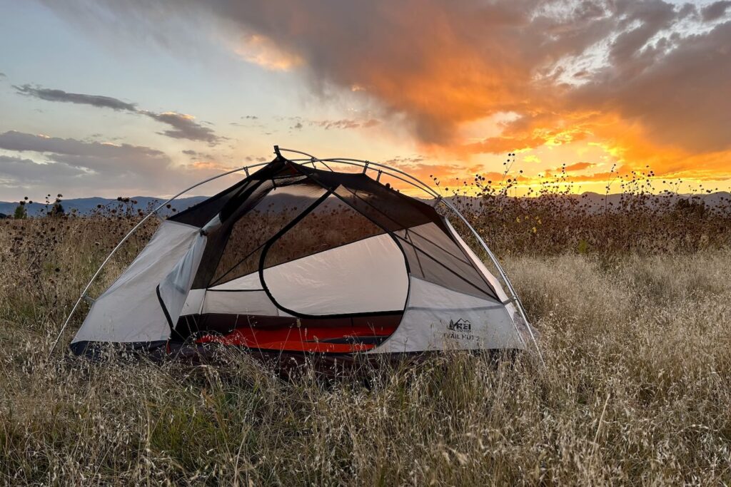 A tent with no fly on sits in a grassy field with a sunset in the background.