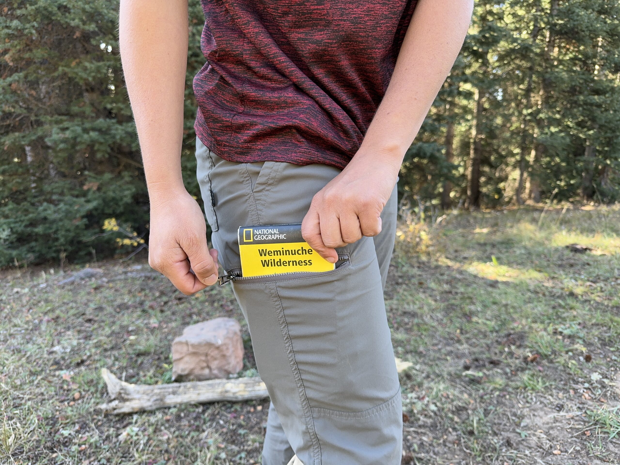 a hiker putting a folded waterproof paper map in the zipper pocket of their rei trailmade pants