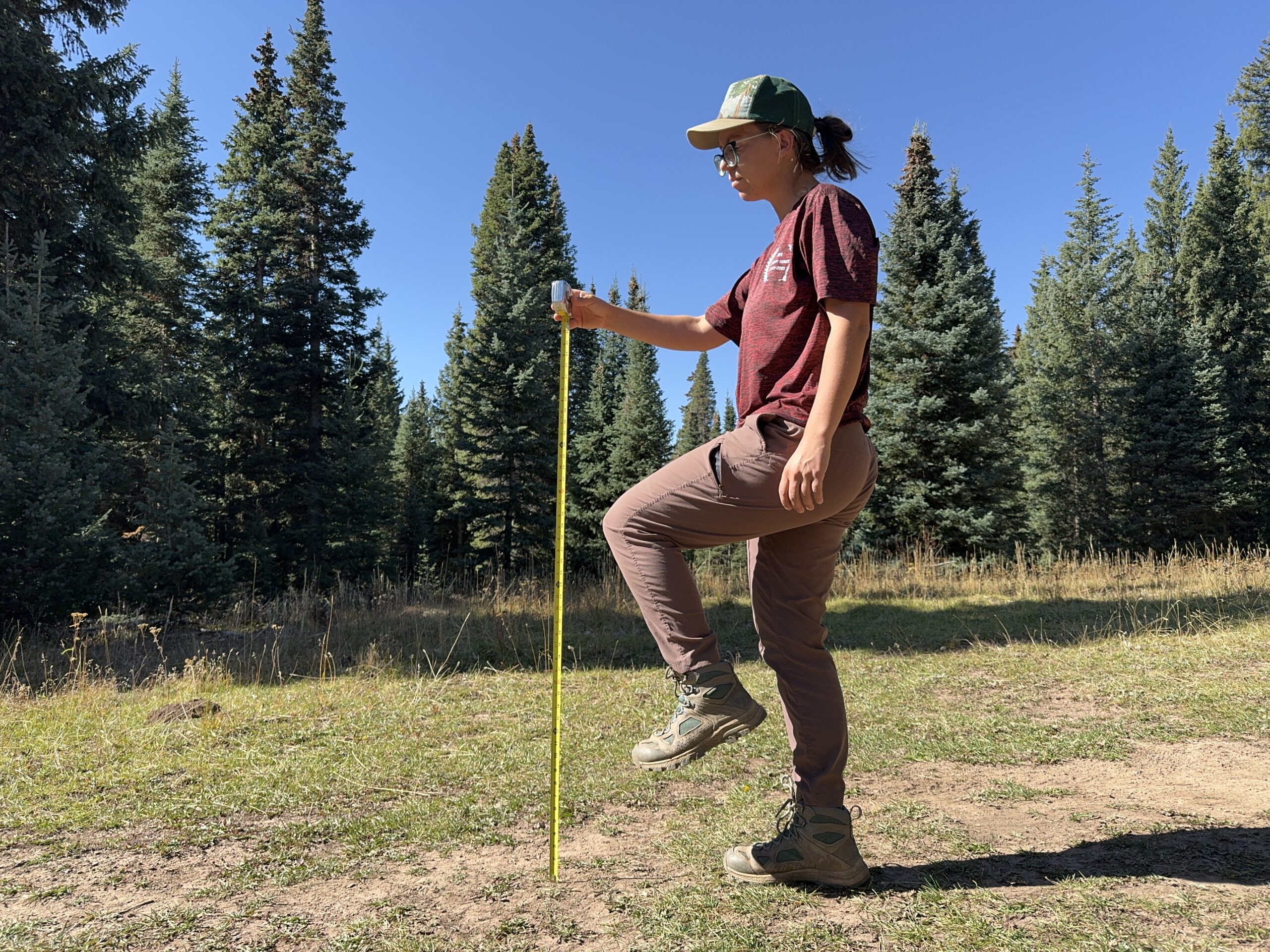 A person is standing with one leg raised in the air while holding a tape measure next to the leg. They are wearing the Mountain Hardwear Dynama pants.