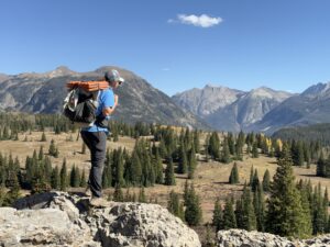 a backpacker on a ledge looking into a forested meadow in the mountains