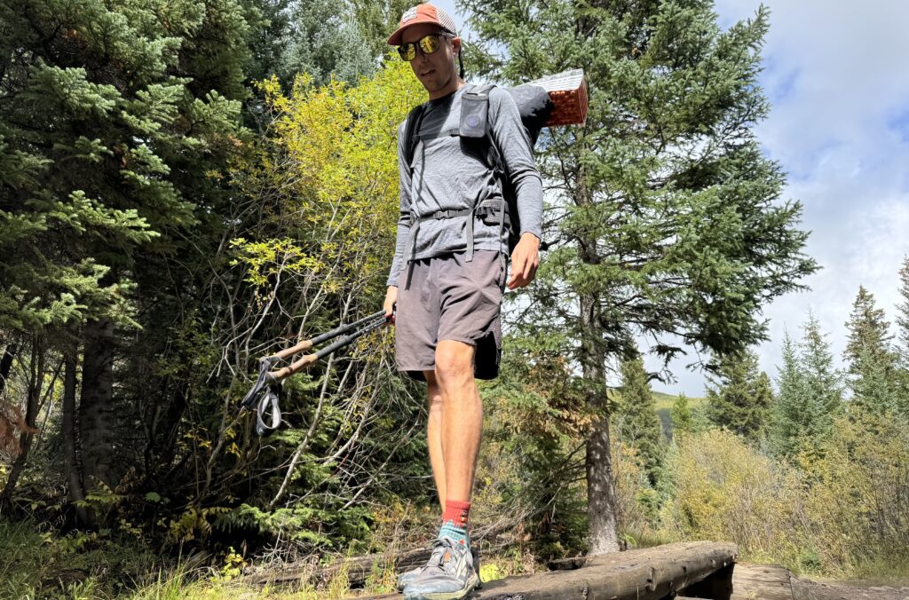 A hiker crosses a log wearing a backpack and the Smartwool Classic All-Season base layer. Trees are in the background.