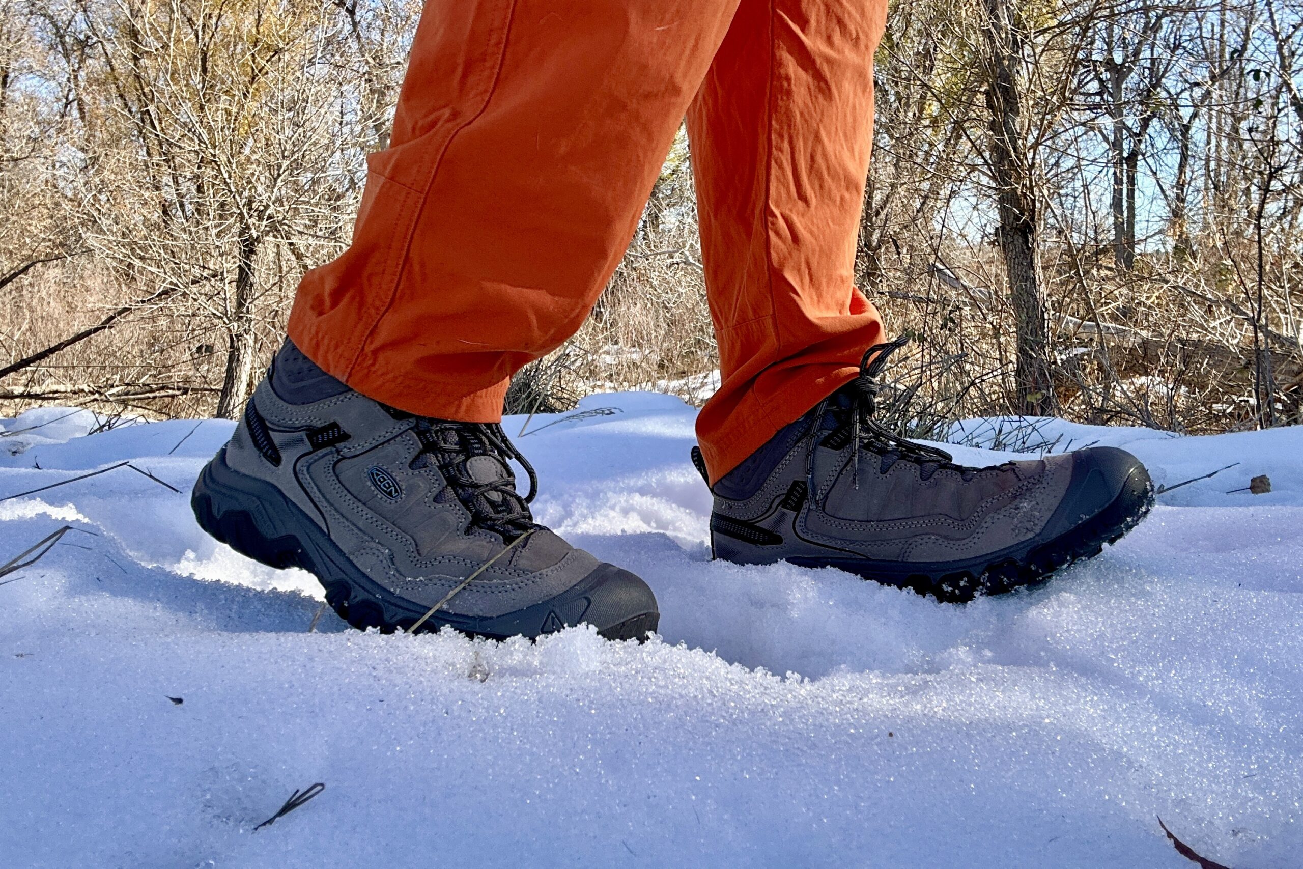 Close up image from knees down of a person wearing hiking boots and bright orange pants walking through snow.