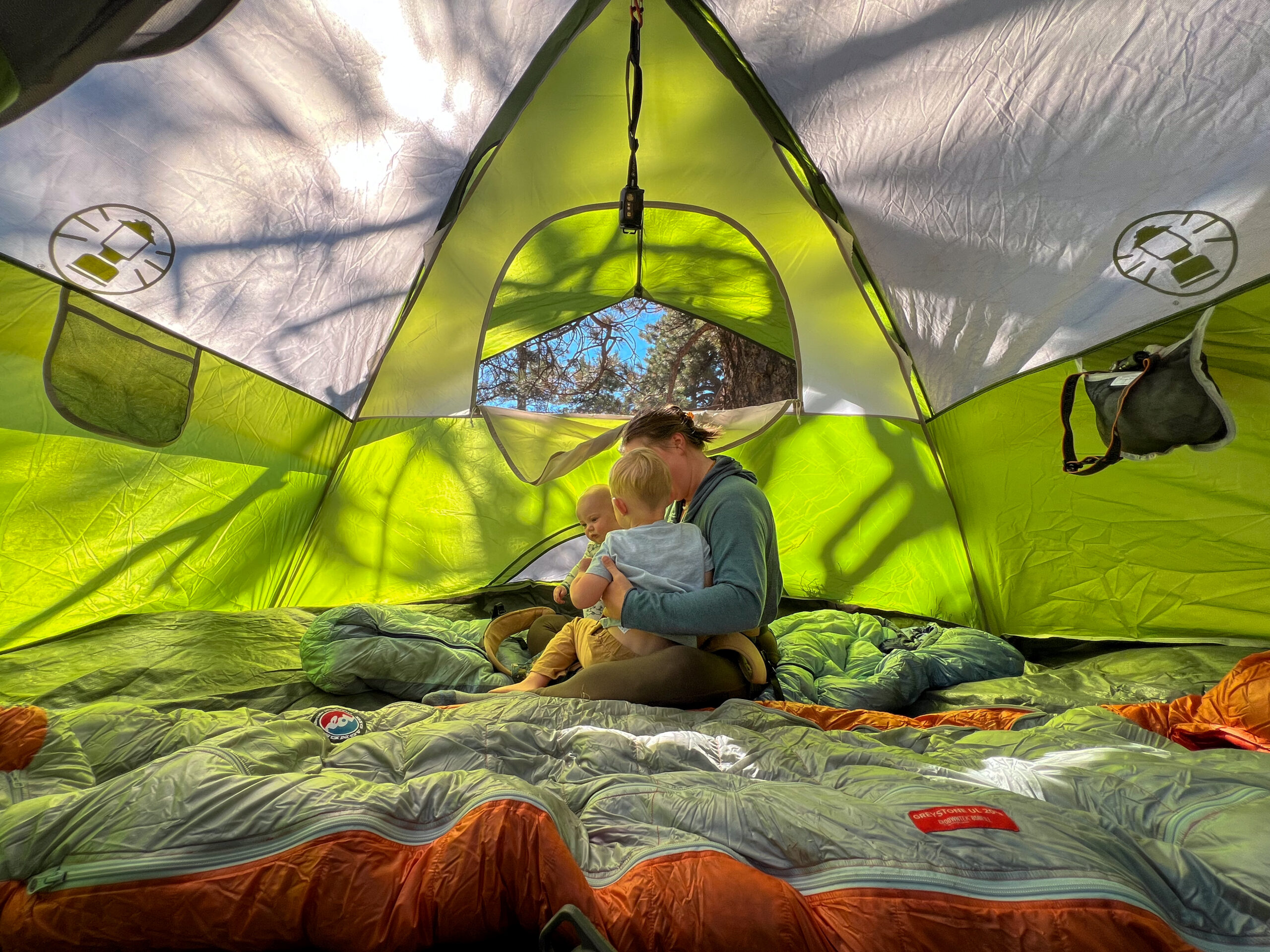 A parent holding a young child inside the Coleman Sundome 4 tent. The tent is spacious, with sleeping bags spread out on the ground. The interior is brightly lit with sunlight streaming through the tent’s fabric, highlighting the green and white colors of the tent. The family is sitting on the bedding, with camping gear visible in the background.