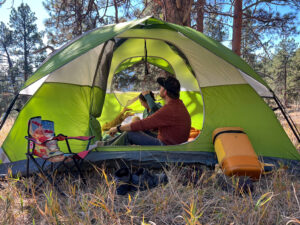 A person sitting inside the Coleman Sundome 4 tent with a young child, both facing toward the front of the tent. The person is wearing a red shirt and holding a snack bag, while the child is resting on the ground. Outside the tent, you can see trees and sunlight filtering through. A camping chair and a yellow cooler are positioned outside the tent.