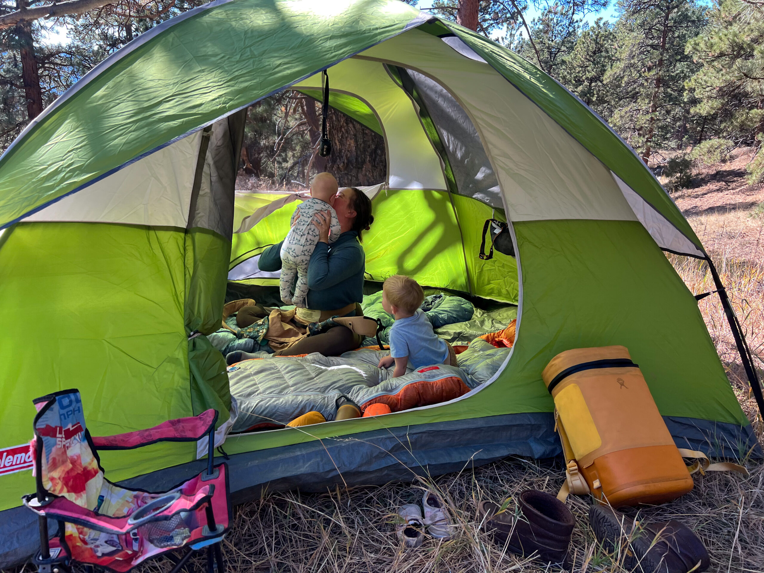 A family of three is inside the Coleman Sundome 4 tent, with one child sitting on the tent floor and the other being held by the mother. The tent is set up in a forested area, with a camp chair, shoes, and a cooler placed outside the tent.