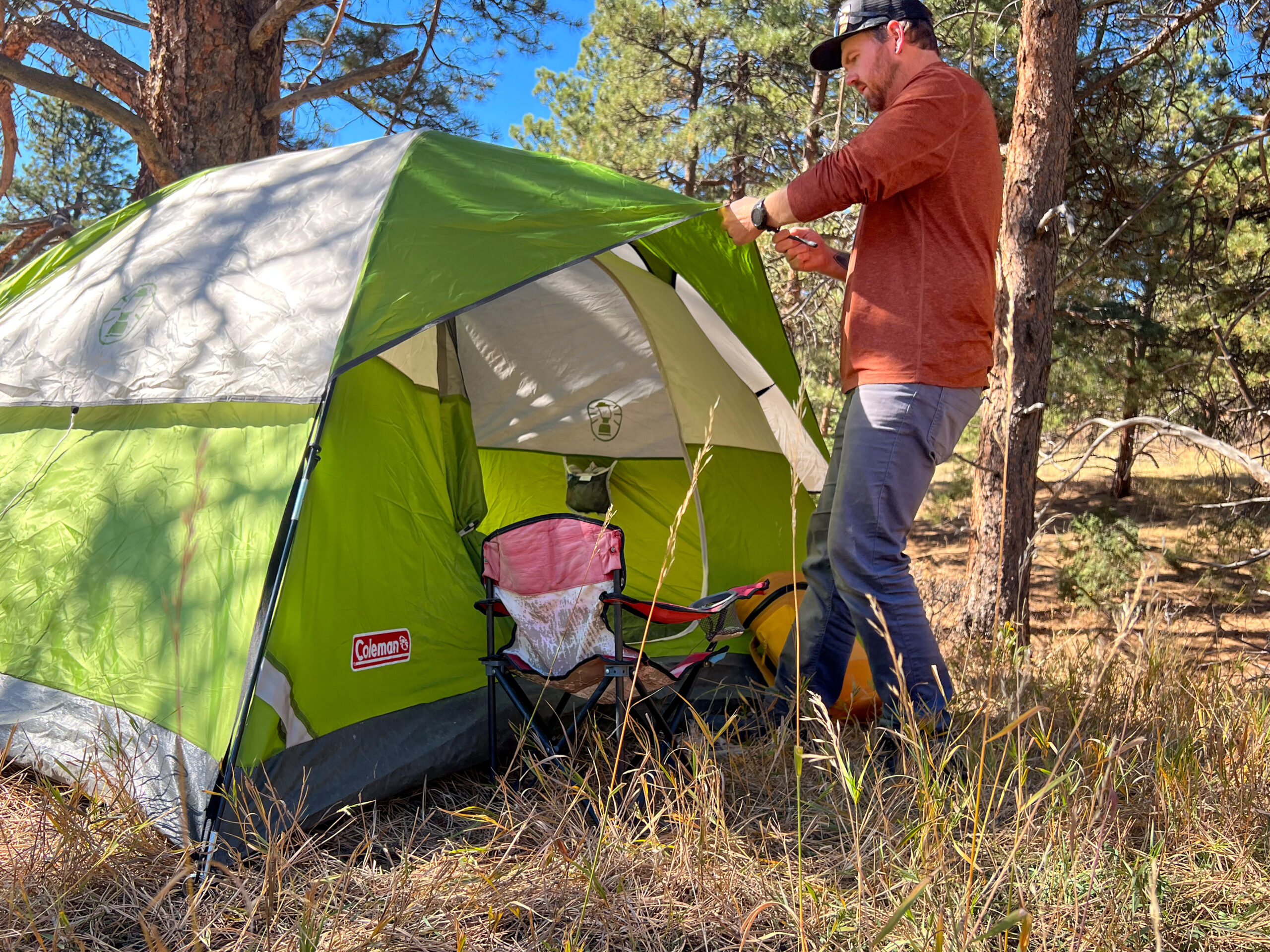 A person is attaching the rainfly to the Coleman Sundome 4 tent, which is set up in a forested area. The tent is green with gray and white panels, and a small pink camping chair is visible near the entrance. The person is wearing a red shirt and gray pants, and the background shows trees and dry grass.