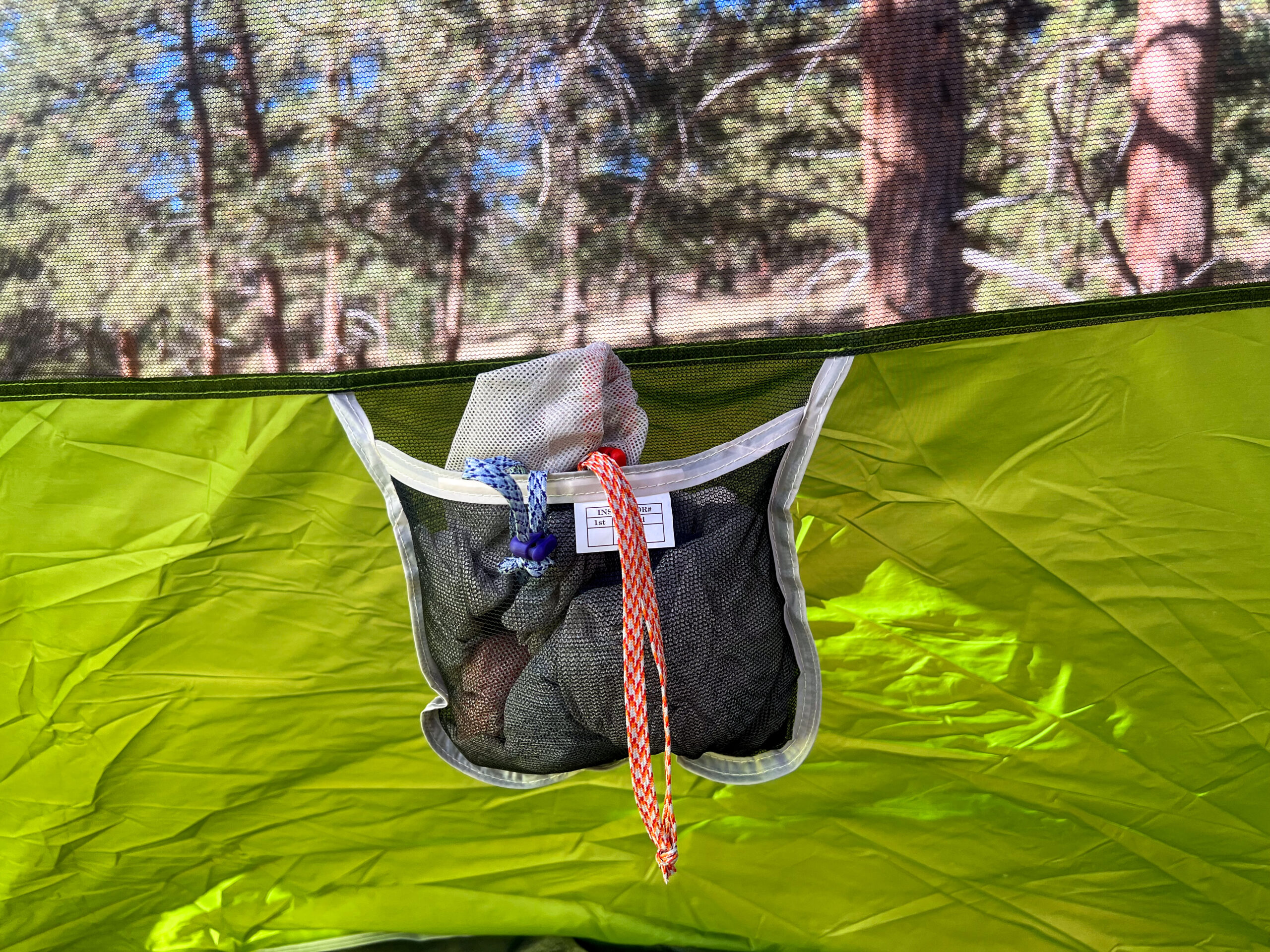 A close-up of a storage pocket inside the Coleman Sundome 4 tent, showing a mesh bag hanging from the tent's interior. The pocket contains a few small items, and the green fabric of the tent is visible surrounding it. The outdoor forest is seen through the tent's mesh window.