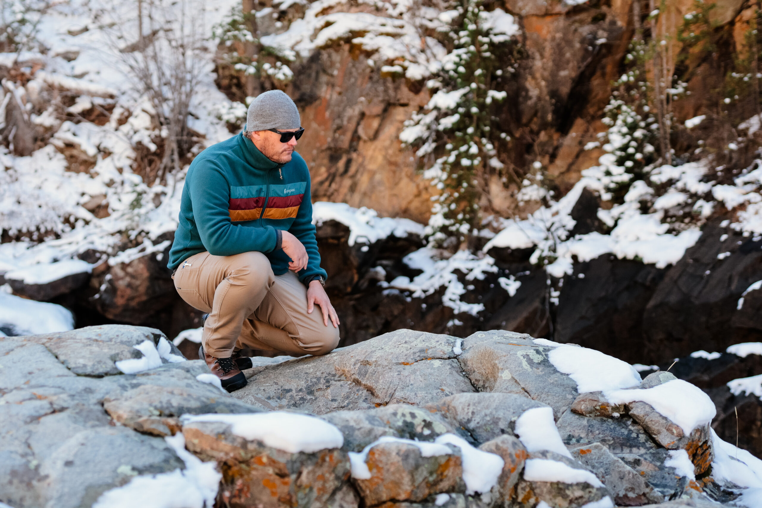 A man kneels on a snowy rock point overlooking the river.