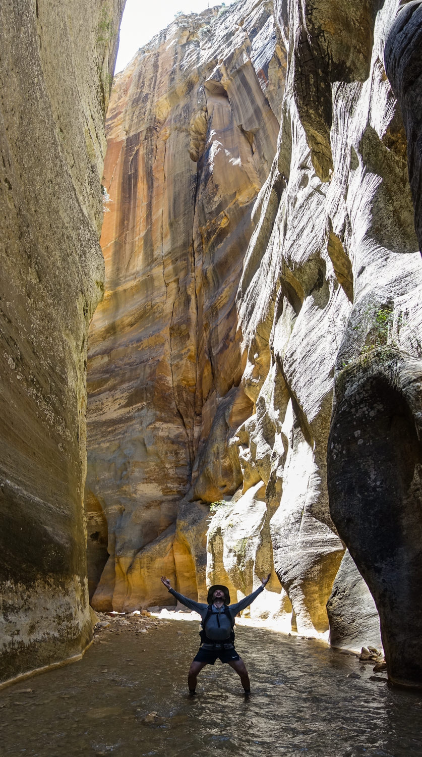 a hiker raises their hands and bends their knees to show their awe as they stand below towering narrow cliffs along the north fork of the virgin river in zion national park